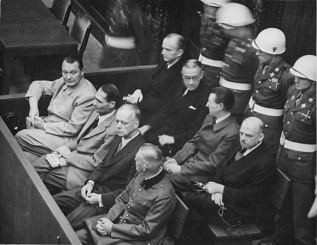 Zdjęcie przedstawia grupę mężczyzn siedzących przy drewnianych ławach. Za nimi stoją żołnierze.