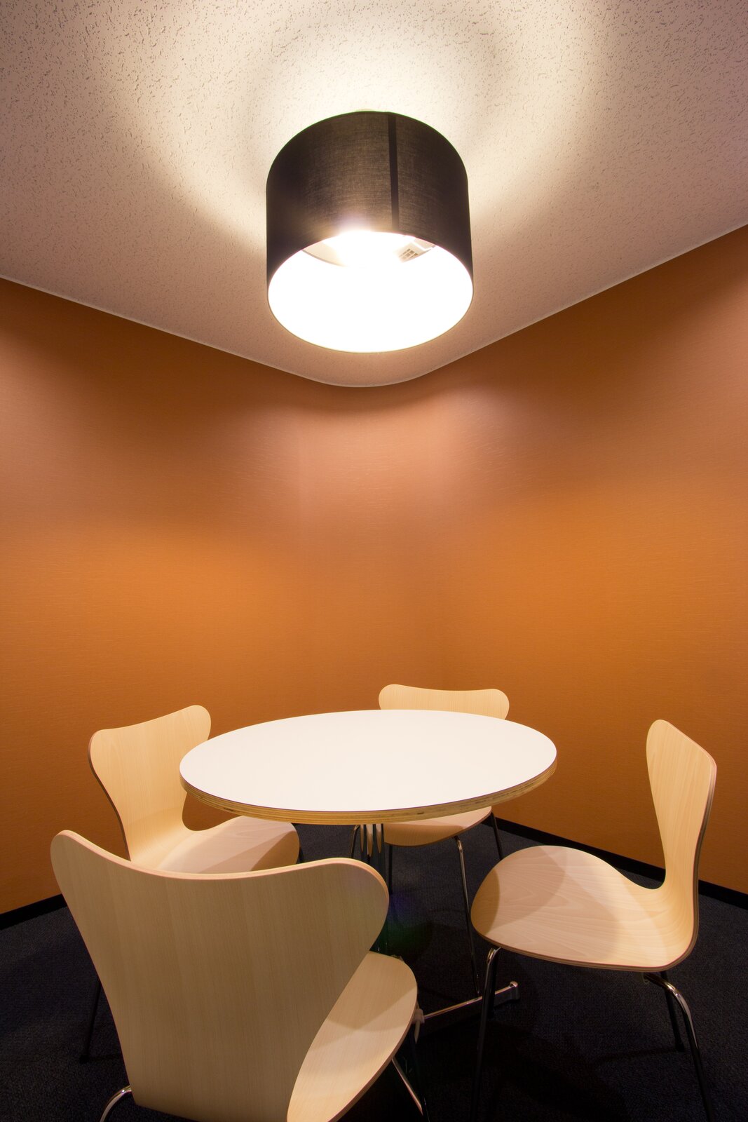 Ilustracja przedstawia pomieszczenie, w którym stoi okrągły, biały stolik i cztery drewniane krzesła. Nad stołem zawieszona jest lampa. Jej światło pada na blat stołu i krzesła. Słabiej oświetla brązową ścianę oraz fragment sufitu.