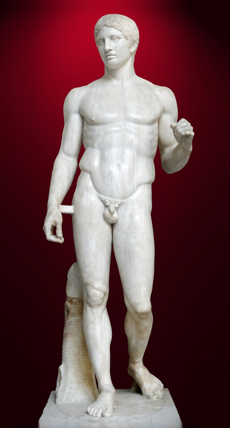 Zdjęcie przedstawia marmurową rzeźbę młodego, nagiego mężczyzny. Ma krótkie, układające się włosy, jest atletycznie zbudowany. Lewą rękę ma nieznacznie uniesioną.