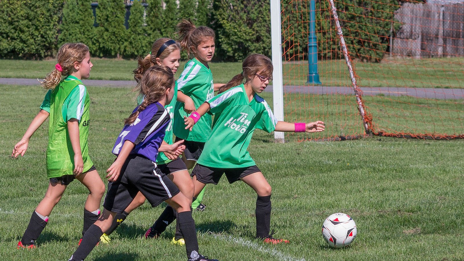 Zdjęcie przedstawia pięć dziewczynek grających w piłkę nożną na murawie. Wszystkie ubrane są w stroje piłkarskie. Trzy z nich w zielone koszulki i czarne szorty, jedna z tyłu w jasnozieloną koszulkę i czarne szorty, a piąta w strój bramkarza. Dziewczynka najbardziej wysunięta do przodu zamierza się do kopnięcia piłki. Po prawej stronie widoczna jest bramka.