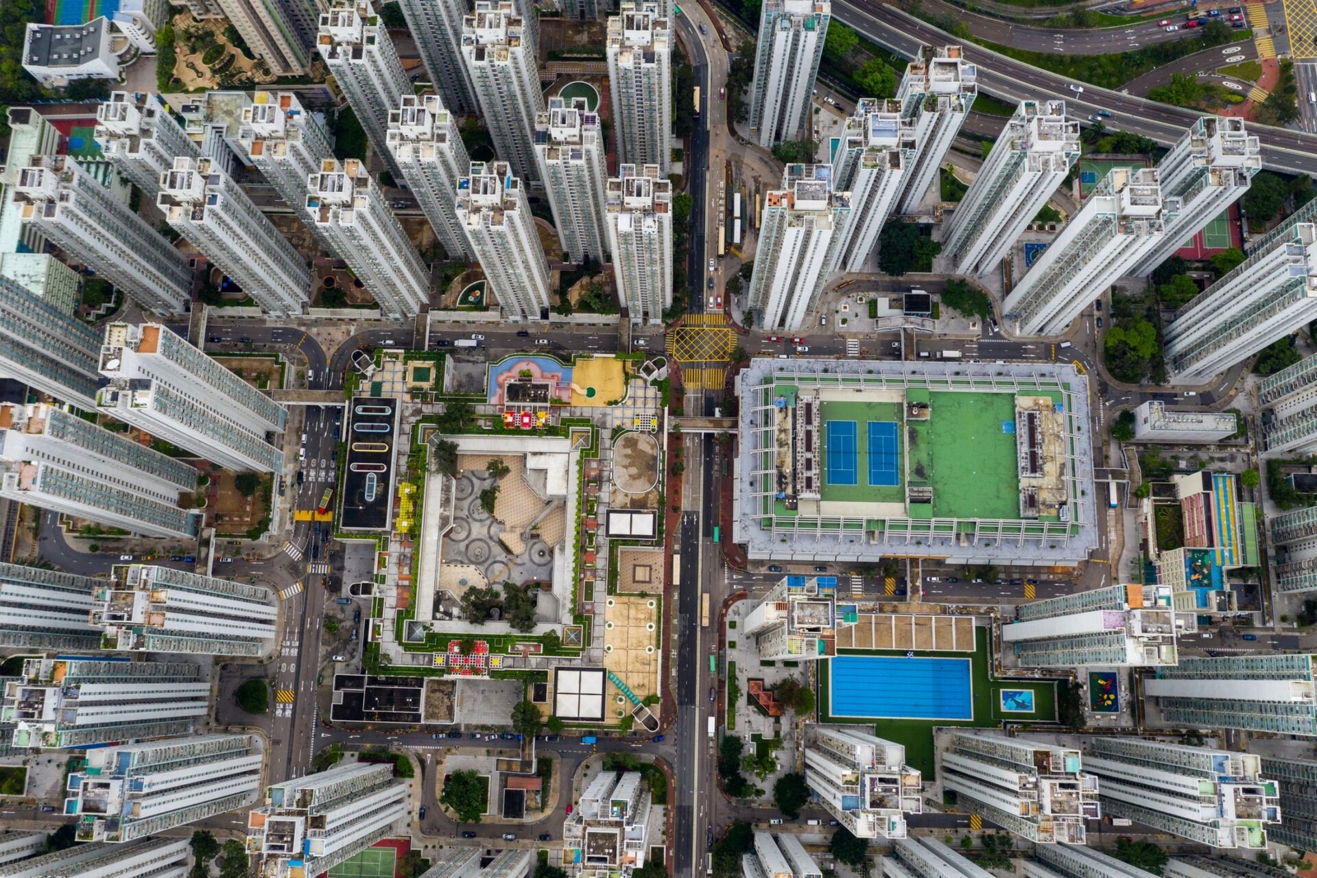 Fotografia wykonana z perspektywy lotu ptaka ukazuje osiedle wieżowców zbudowane dookoła prostokątnego terenu  rekreacyjnego, który przecina trasa komunikacyjna.