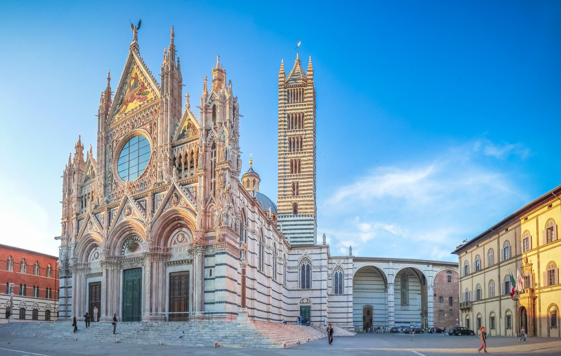 Ilustracja o kształcie poziomego prostokąta przedstawia fotografię Katedry w Sienie. Widok ukazuje katedrę od strony zachodnio-południowej. W portalu zachodnim znajdują się trzy wejścia z półokrągłym tympanonem, otoczonym archiwoltami i zwieńczonym trójkątnym przyczółkiem.  Ściana frontowa zamknięta jest od góry trójkątnym zwieńczeniem z dekoracją rzeźbiarską i wieżyczkami, znajdującymi się po bokach. Wieżyczki znajdują się także na obrzeżach. W fasadzie widoczne są poziome podziały ściany. Po prawej stronie ukazany jest fragment transeptu oraz dwie wysokie, kwadratowe wieże. Do kościoła dostawiony jest budynek z arkadami. Po bokach kościoła widoczna jest zabudowa miejska.