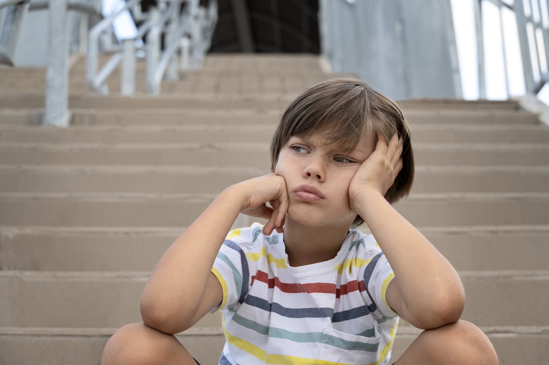 Zdjęcie przedstawia chłopca siedzącego stopniach wysokich schodów. Chłopiec ma głowę skierowaną lekko w lewą stronę i patrzy w lewo. Ma ugięte nogi. Ręce ma zgięte w łokciach i oparte na kolanach. Prawa dłoń ma podwinięte palce i dotyka prawego policzka, zaś lewa ma palce wyprostowane i dotyka lewej strony twarzy. Chłopiec opiera na niej głowę. Ubrany jest w bluzkę z krótkim rękawem w kolorowe paski na białym tle. Ma krótkie, ciemne włosy. W tle widoczne stopnie schodów oraz srebrne poręcze.  