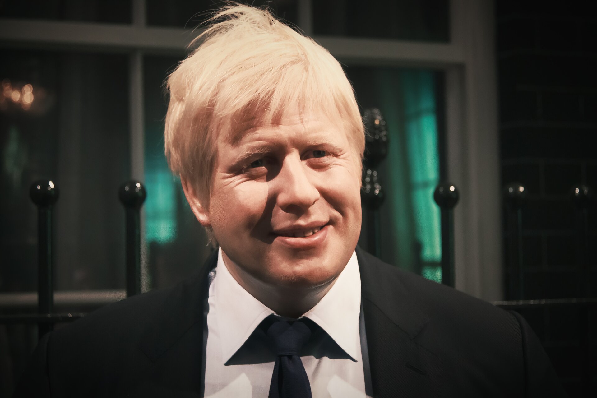 Woskowa podobizna Boris Johnson. Zdjęcie ukazuje postać mężczyzny w średnim wieku od klatki piersiowej w górę. Mężczyzna ma dłuższe, blond włosy, okrągłą twarz i jasną karnację. Ubrany jest w czarny garnitur, białą koszulę i czarny krawat.