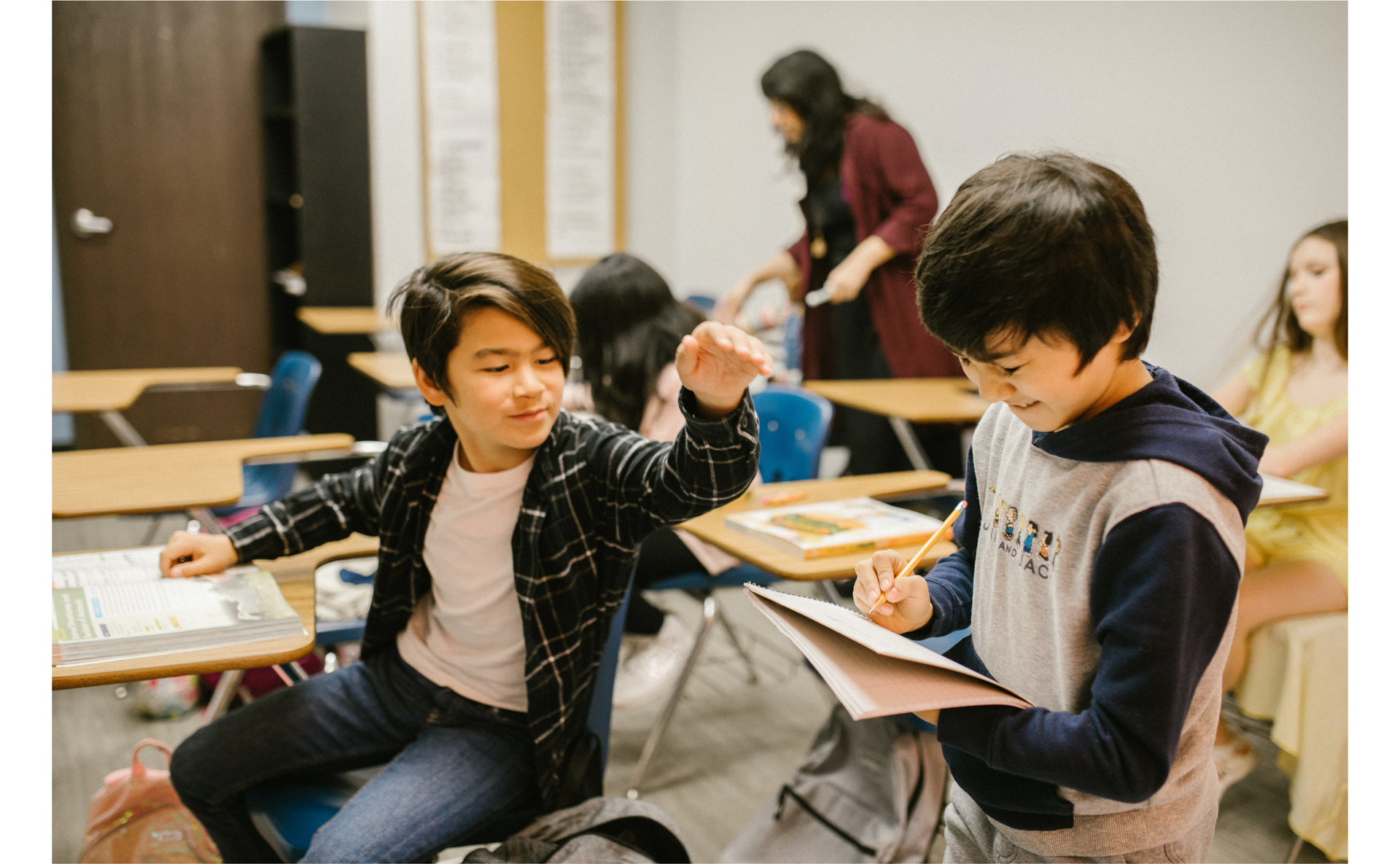 Zdjęcie przedstawia dwóch chłopców w klasie szkolnej. W tle widoczne są inne osoby. Chłopiec po lewej stronie wyciąga rękę w stronę chłopca znajdującego się po prawej stronie zdjęcia. Chłopiec po prawej trzyma w ręku jakiś notes i ołówek.