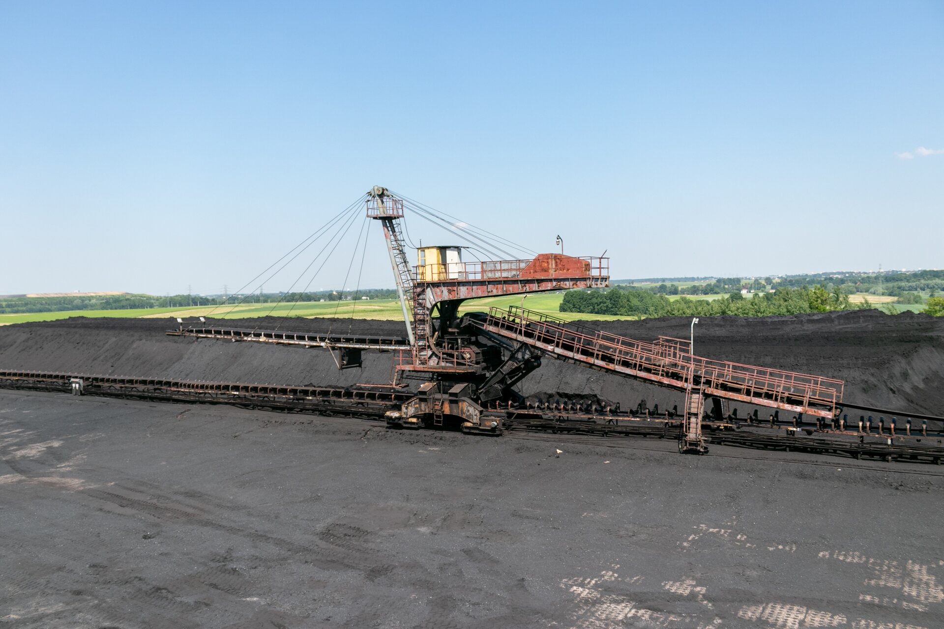 Na zdjęciu widoczna jest zwałowarka. Zwałowarka jest maszyną wielkogabarytową. Z prawej strony do zwałowarki dołączony jest przenośnik taśmowy. Z przodu zwałowarki znajduje się drugi przenośnik taśmowy. Przenośnik jest odchylony od poziomu. Przenośnik podwieszony jest na linach. Liny zamocowane są na głównym przęśle. Z przenośnika spada zrzucany na hałdę węgiel. W tle widać dużą hałdę węgla.Poniżej zdjęcia widoczny jest przycisk multimedialny z nagraniem audio. Treść nagrania jest zgodna z tytułem zdjęcia.