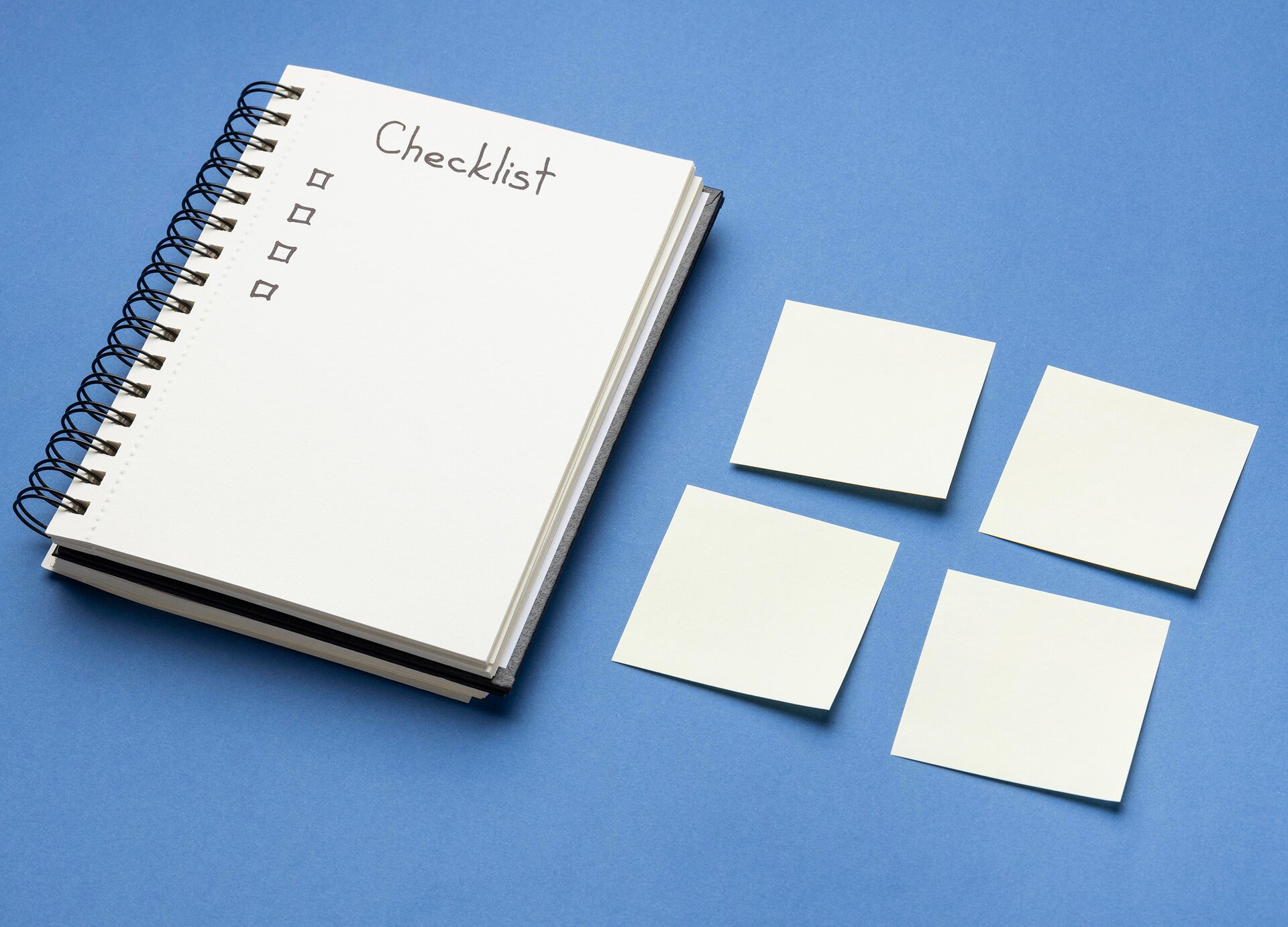 Zdjęcie przedstawia otwarty notes z grzbietem zszytym czarną spiralą. Na otwartej stronie widoczny jest napis w języku angielskim: CHECKLIST, poniżej którego widoczne są cztery odręcznie narysowane kwadraciki. Na prawo od notesu widoczne są cztery samoprzylepne karteczki. 