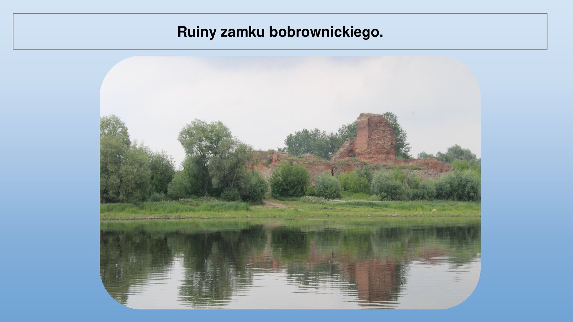 Niebieski slajd. U góry nagłówek: „Ruiny zamku bobrownickiego”. Niżej zdjęcie przedstawiające ruiny zamku. Widoczny jest fragment muru i wieży z prawej strony. Zamek zbudowany jest z czerwonej cegły. Przed ruinami zielone drzewa, a dalej, u dołu zdjęcia, niebieska woda.