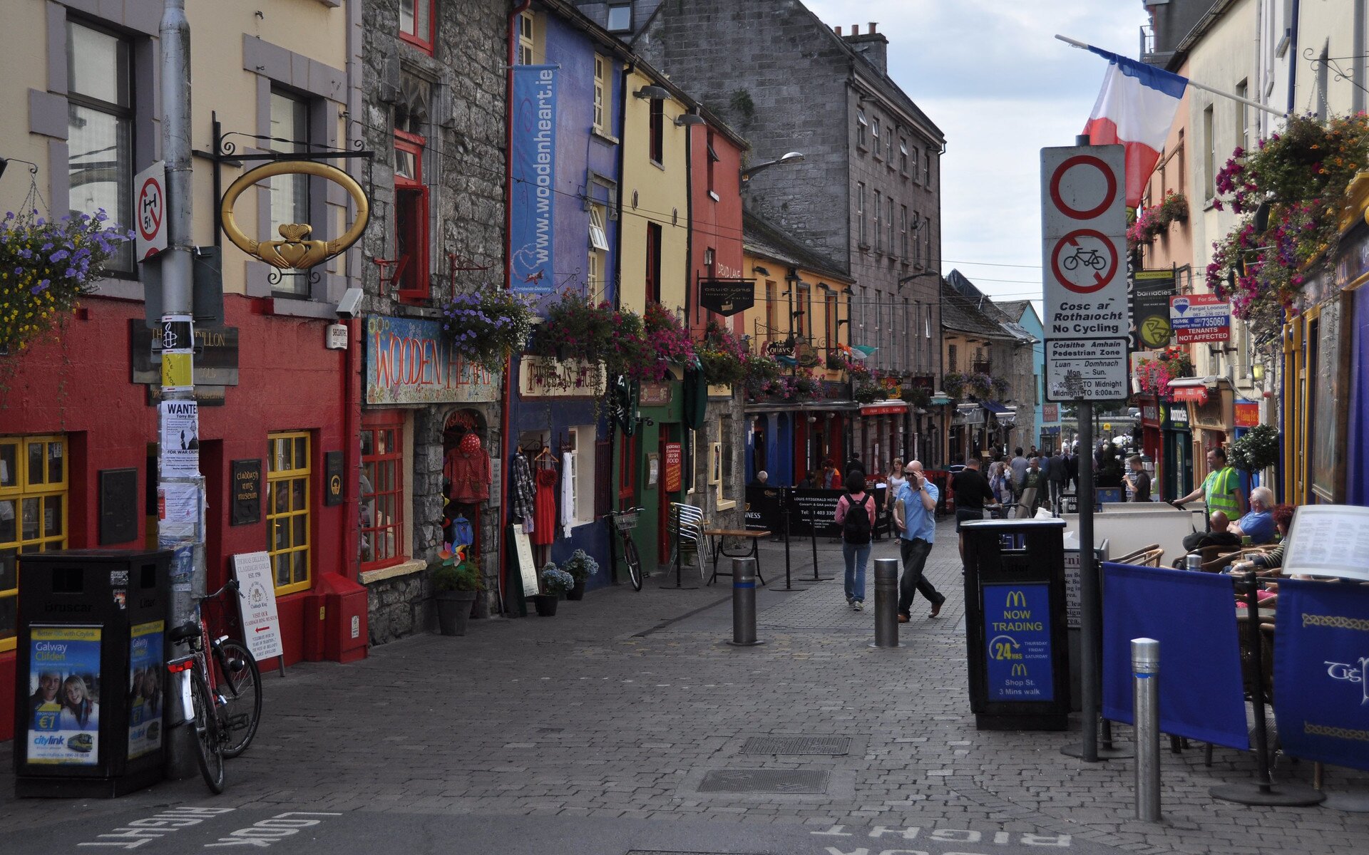 Zdjęcie przedstawia niewielką uliczkę, wzdłuż której, po obu stronach, stoją domki z elewacjami w różnych kolorach i stylach architektonicznych. Na frontach budynków widoczne są szyldy sklepowe, powieszone są kwiaty w doniczkach, wystawione ubrania. W tle widoczna jest markiza, pod którą stoją stoliki i krzesła. Po prawej stronie widoczne są krzesła i stoliki przy których siedzą ludzie. Obok widoczny jest znak zakazu wjazdu z napisem: No Cycling, Pedestrian Zone.