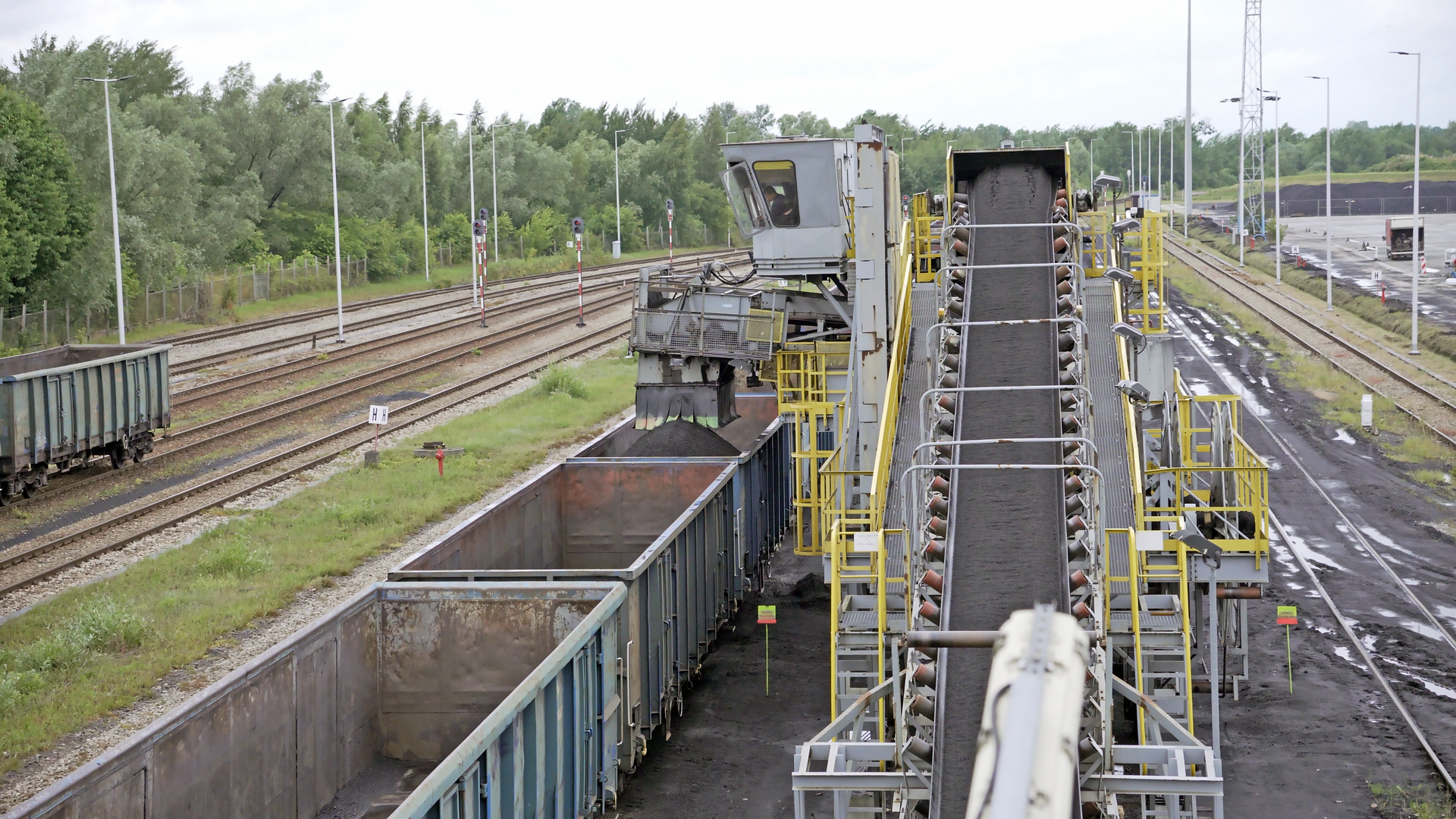 Na zdjęciu widać proces napełniania wagonów węglem. Pierwsze dwa wagonu są puste. Ostatni wagon jest napełniany. Nad wagonem jest nasypywarka. Nasypywarka przypomina prostopadłościan z którego wysypuje się bezpośrednio do wagonu węgiel. Nad nasypywarkom znajduje się kabina operatora. Równolegle do składu biegnie taśmociąg. Na taśmie taśmociągu transportowany jest węgiel.Poniżej zdjęcia widoczny jest przycisk multimedialny z nagraniem audio. Treść nagrania jest zgodna z tytułem zdjęcia.