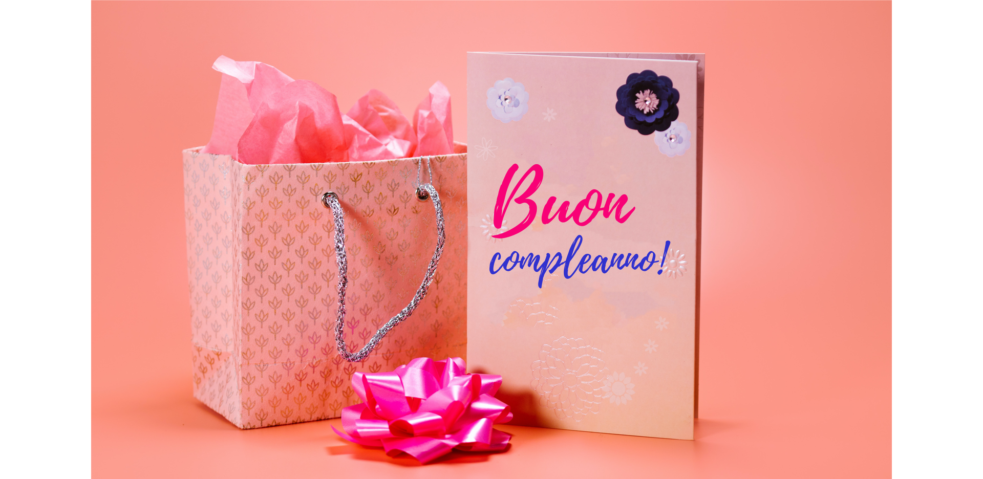 Na zdjęciu po lewej stronie stoi torba z prezentem. Po prawej znajduje się kartka w kwiatki z napisem „Buon compleanno!”. Po środku leży duża, papierowa, błyszcząca kokarda.