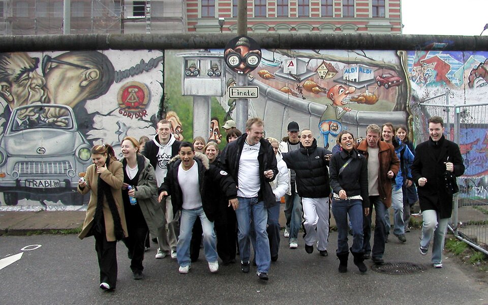 Zdjęcie przedstawia grupę kilkudziesięciorga idących przed siebie ludzi. Są oni uśmiechnięci. Za ich plecami rozciąga się mur z namalowanym graffiti. Jest to fragment Muru Berlińskiego - zabytku związanego z historią Niemiec.