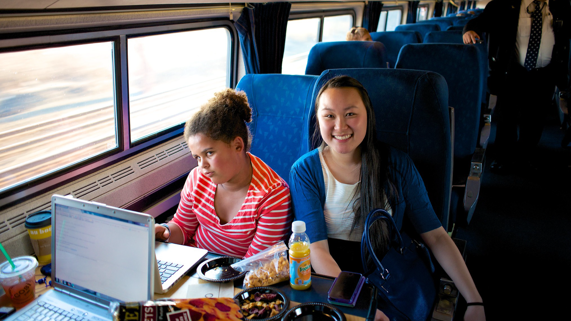 Zdjęcie przedstawia dwie dziewczyny, które siedzą w fotelach w pociągu. Jedna z dziewczyn szeroko się uśmiecha. Przed nimi na stolikach znajdują się laptopy, coś do jedzenia, napój oraz telefon.