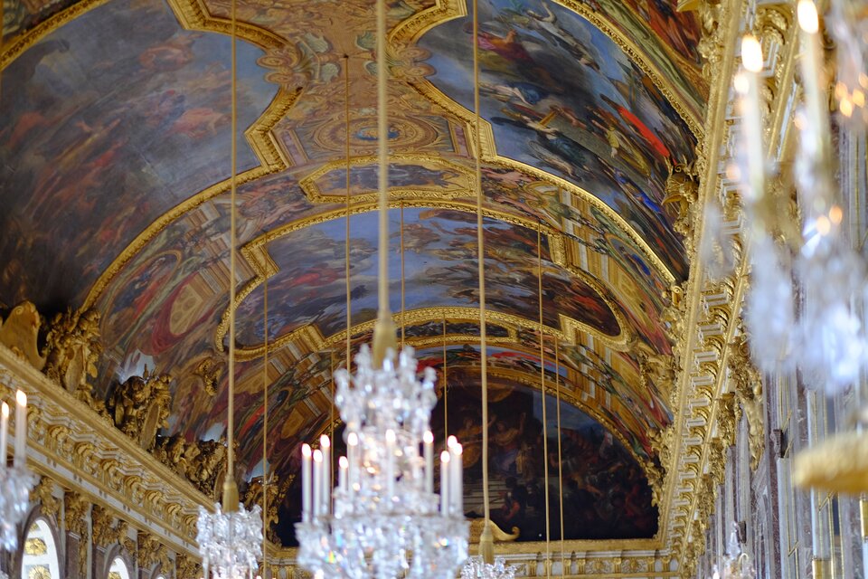 Zdjęcie przedstawia sufit w Sali Luster w Wersalu. Na suficie znajdują się ozdobne plafony w złotych ramach, przestawiające sceny mitologiczne. Na ścianach przy suficie znajdują się rzeźbione złote gzymsy i pilastry ze złotymi ozdobami. Z sufitu zwisają kryształowe żyrandole. 