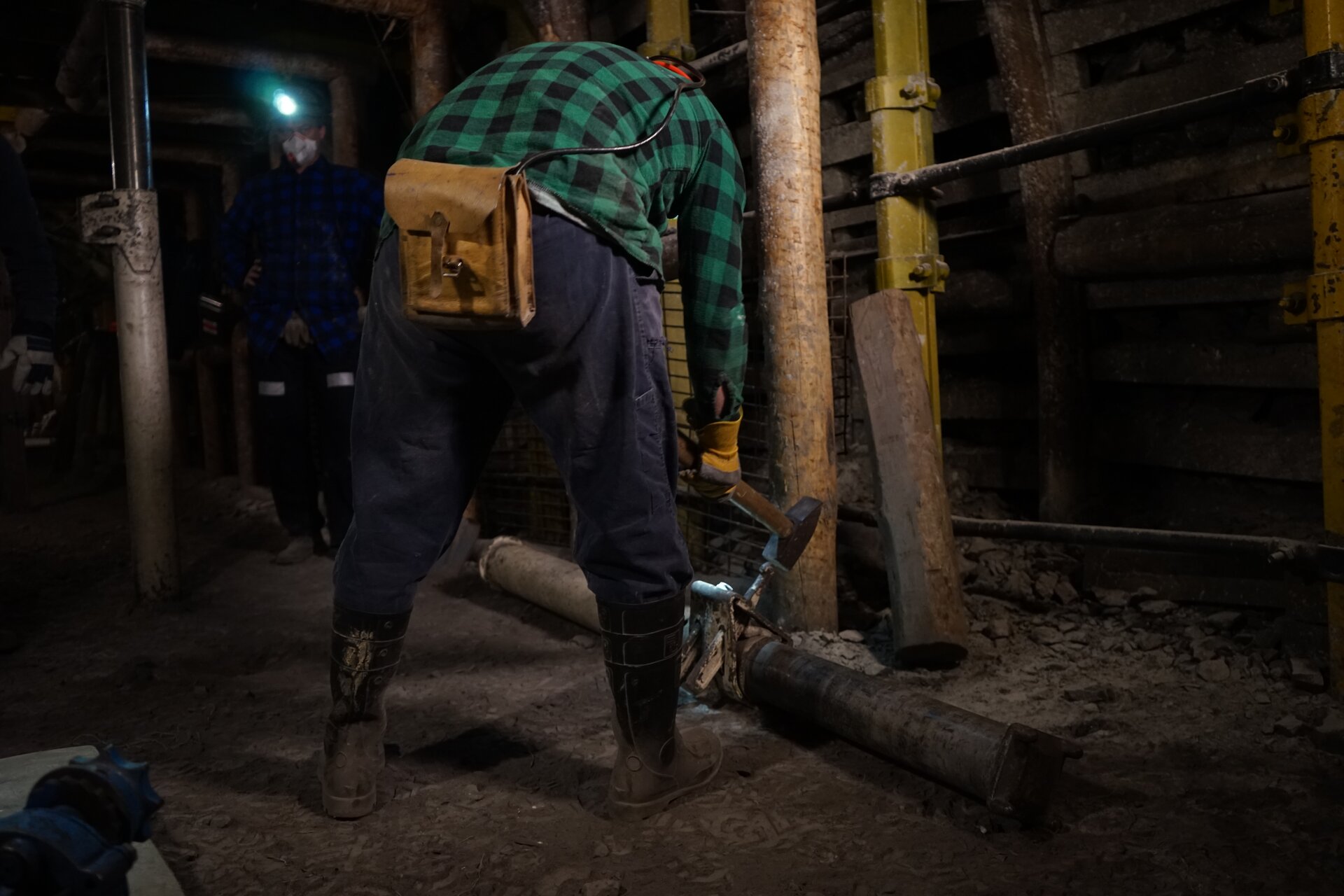 Na zdjęciu widać górnika trzymającego młot górniczy. W centralnej części zdjęcia znajduje się górnik ubrany w strój roboczy. W rękach ma młot. Młot ma drewniany człon. Obuch jest stalowy. Młotek jest zbliżony kształtem do litery T. Górnik uderza młotkiem o rurę znajdując się na spągu. Rura jest najpewniej fragmentem lutniociągu. W tle widać obudowę górniczą.