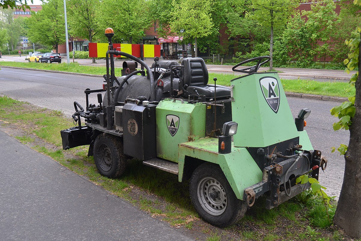 Zdjęcie przedstawia skarpiarkę podpoziomową. Widoczne jest urządzenie w ujęciu z boku. W przedniej części znajduje się miejsce operatora. W tylnej części widoczne są zbiorniki na niewielkiej platformie, połączone rurkami, z zaworami, kablami. Skarpiarka stoi na pasie zieleni między drogą a chodnikiem. W tle widoczna ulica.

Opisy obiektów prezentowanych na grafikach są załączone w treści galerii zdjęć.