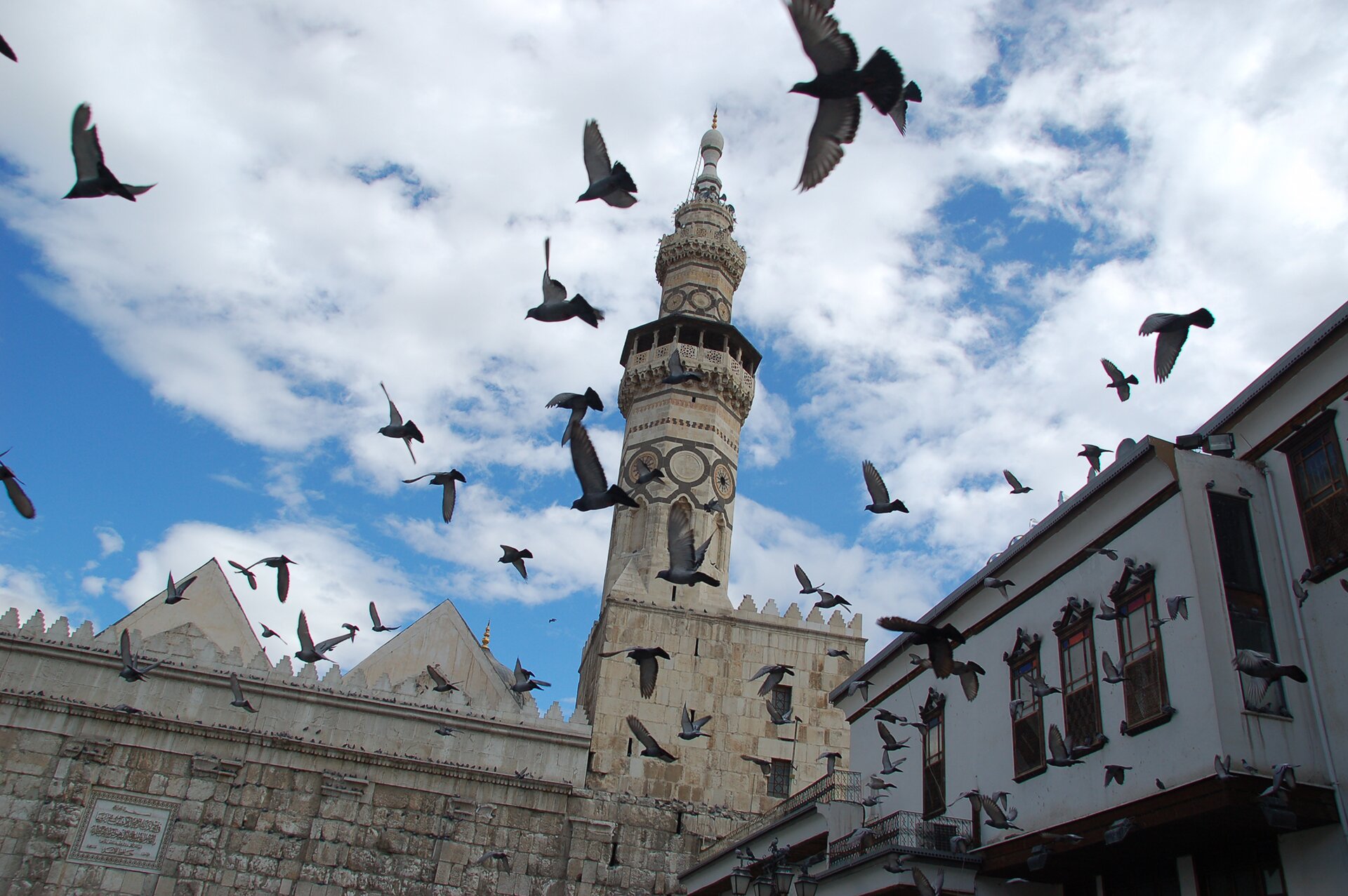 Kliknij, aby powiekszyć. Kolorowej zdjęcie przedstawia minaret i fragment murów wykonanych z jasnego kamienia. Wieża jest ozdobiona malowidłami. Widać przelatująca chmarę czarnych ptaków. Na niebieskim niebie widnieją białe chmury.