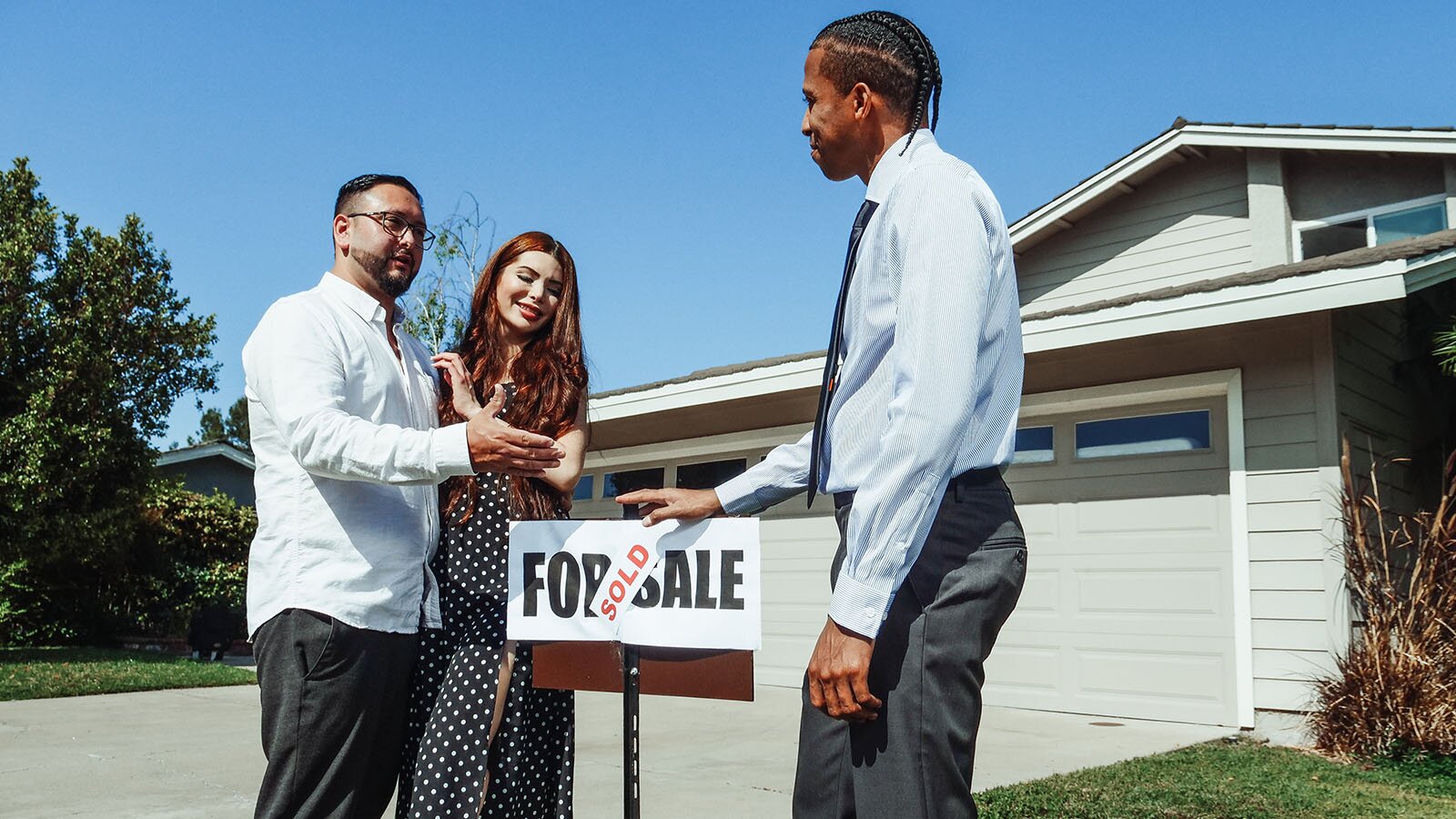 Zdjęcie przedstawia troje osób stojących przy znaku ustawionego przed domem jednorodzinnymFor Sale , na którym jest nalepka Sold. Dwie osoby stoją po lewej stronie znaku. Mężczyzna wyciąga przed siebie rękę, a kobieta jest po jego lewej stronie. Naprzeciwko po drugiej stronie stoi mężczyzna ubrany w białą koszulę, spodnie od garnituru i krawat. Mężczyzna prawą dłonią opiera się o znak. Wszyscy lekko się uśmiechają.