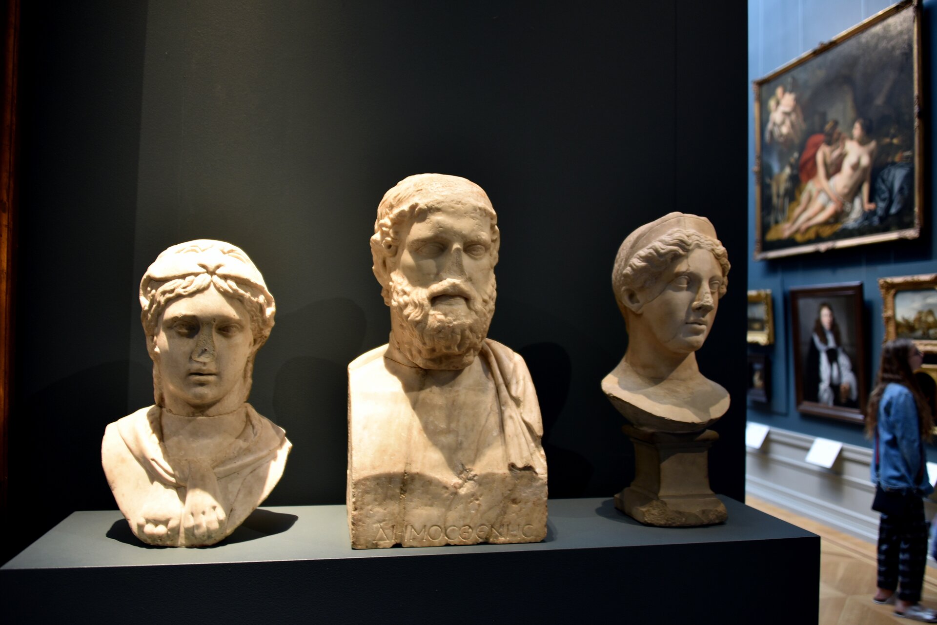 Fotografia przedstawia trzy rzeźby – popiersia dwóch bogiń Diany i Junony i mężczyzny Anakreonta, na tle czarnej ściany oraz muzeum, w którym widoczne są obrazy oraz osoba je obserwująca. Rzeźby wykonane są z jasnego kamienia. Pierwsza z nich przedstawia młodą kobietę z opaską na włosach oraz lekko otwartymi ustami. Jej włosy okalają jej twarz, patrzy ona przed siebie. Druga rzeźba przedstawia starszego mężczyznę o średniej długości zaroście oraz kręconych , krótkich włosach. Trzecia rzeźba ukazuje młodą kobietę z opaską na włosach, której rysy twarzy są delikatne. Nosy dwóch pierwszych rzeźb wydają się ułamane.