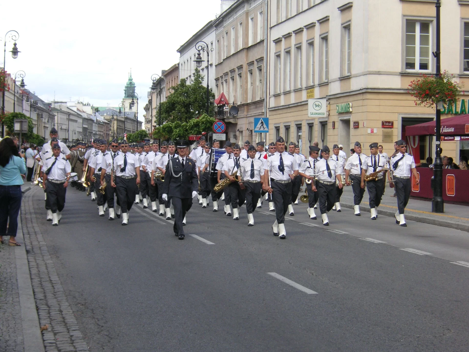 Ilustracja przedstawia orkiestrę dętą typu harmonia. Muzycy ubrani są w czarne spodnie, białe czapki, koszule, pasek i buty. W tle widać kamienice, po lewej stronie na chodniku tłum ludzi.