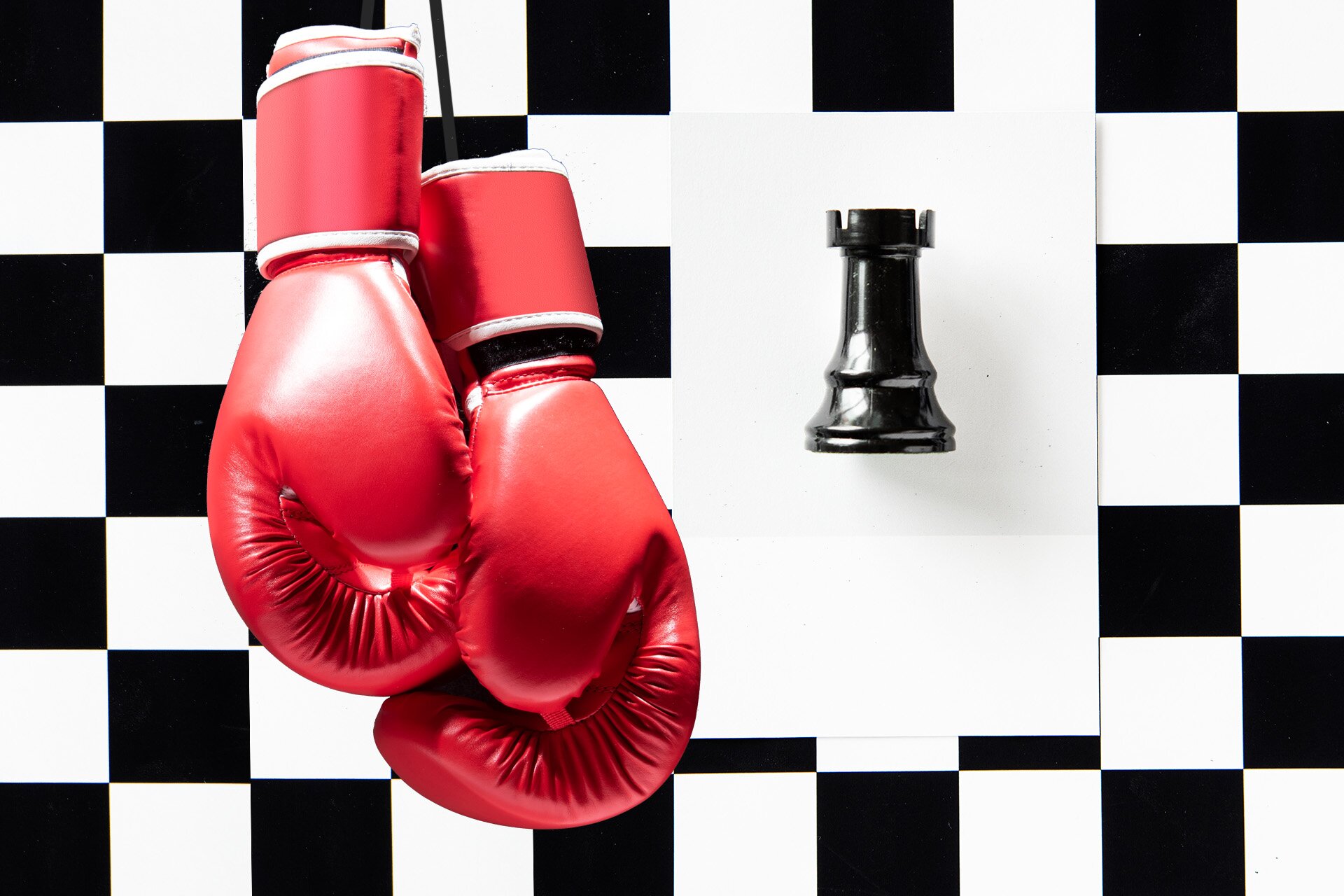 Ilustracja przedstawia wiszące czerwone rękawice bokserskie na tle ściany w szachownicę czarno białą. Obok nich widoczny jest pionek wierzy szachowej. 