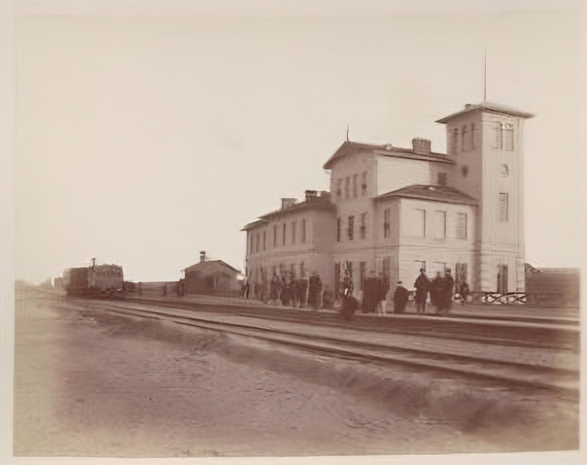 Kliknij, aby powiększyć. Zdjęcie przedstawia budynek stacji (widok z daleka). Na pierwszym planie płaski teren i dalej niezbyt wysoki nasyp kolejowy. Na nasypie dwa rzędy torów. Za torami peron, na którym stoją ludzie. W głębi budynek stacji. Składają się na niego cztery prostokątne połączone części o różnej wysokości. Najwyższa jest trzypiętrowa, narożna wieża. Środkowa część budynku jest nieco niższa, ale również ma trzy kondygnacje. Pozostałe fragmenty budynku stacji są dwukondygnacyjne. W oddali widać inne zabudowania kolejowe.