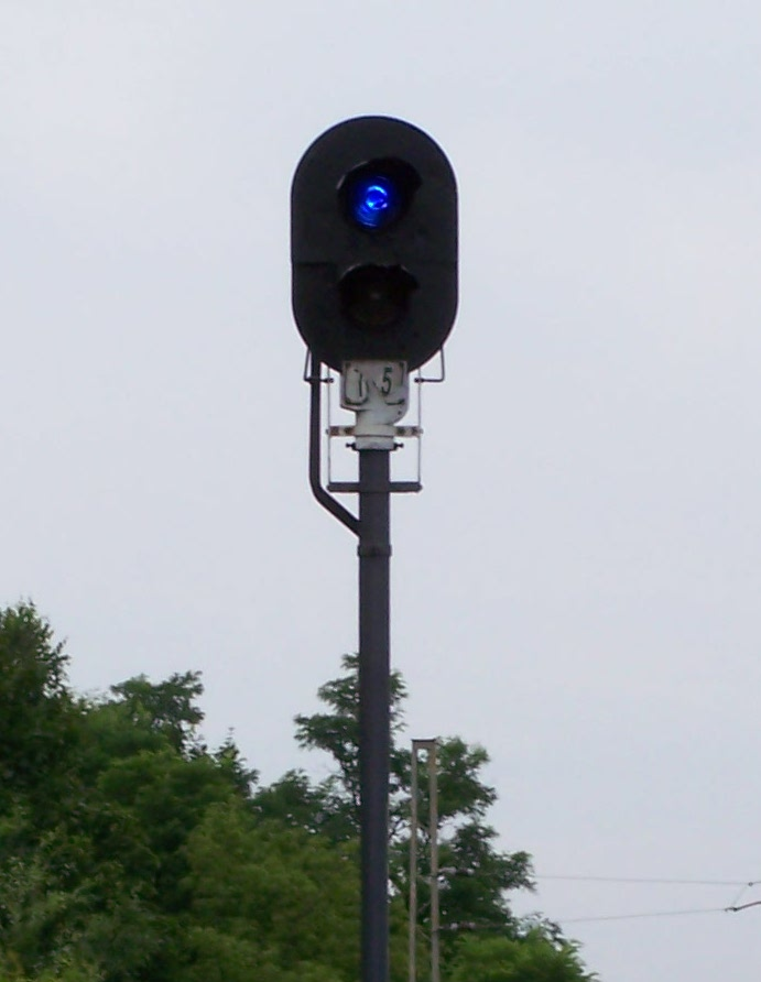 Zdjęcie przedstawia tarczę ostrzegawczą będącą elementem obwodowych świateł tarcz manewrowych. Tarcza znajduje się obok torowiska. Sygnalizator jest umieszczony na szarym maszcie, posiada on dwa światła, przy czym jedno z nich, górne, świeci się na niebiesko. 