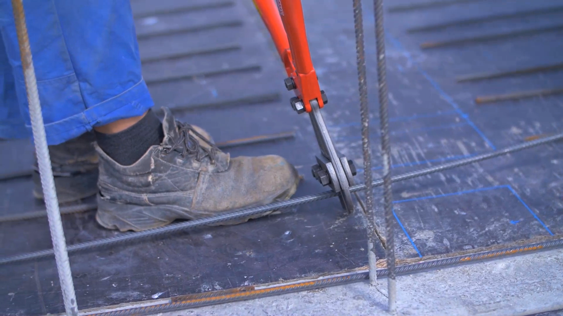 Zdjęcie przedstawia czerwone nożyce zbrojarskie ręczne, za pomocą których robotnik przecina stalowy pręt.