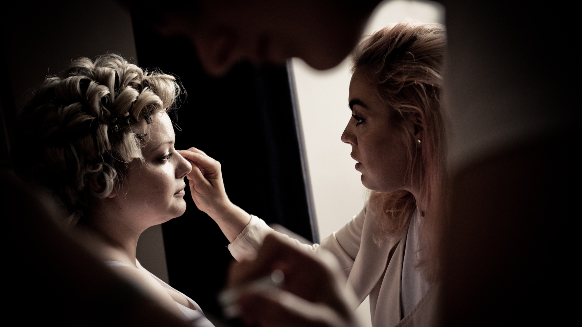 Zdjęcie przedstawia dwie kobiety. Jedna z nich (makijażystka) wykonuje makijaż/maluje drugą kobietę.