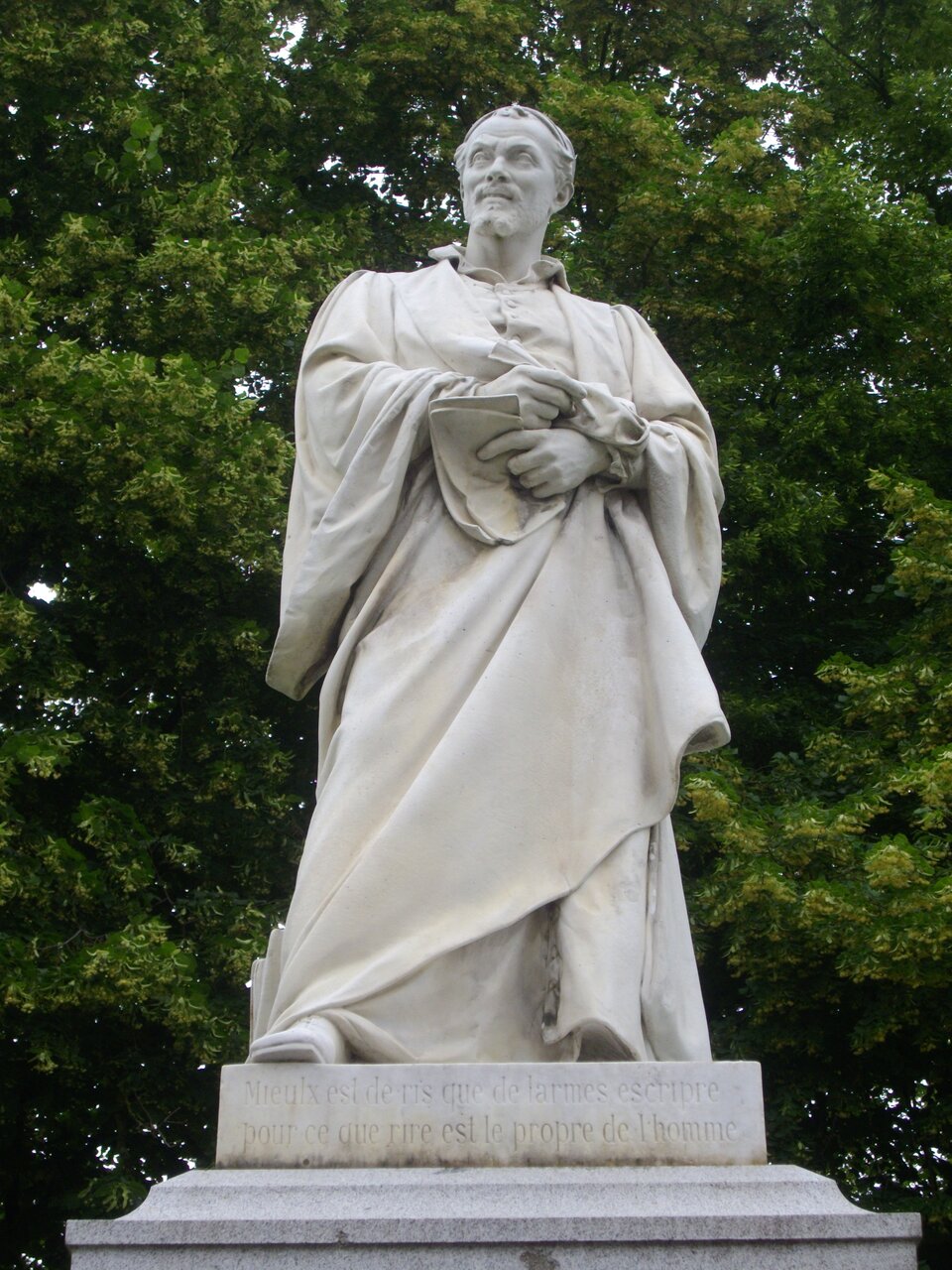 Zdjęcie przedstawia pomnik. Na postumencie znajduje się stojący mężczyzna. Jest dojrzały, ma szczupłą twarz. Ubrany jest w sięgający ziemi płaszcz. Trzyma w lewej ręce kilka kartek papieru. 