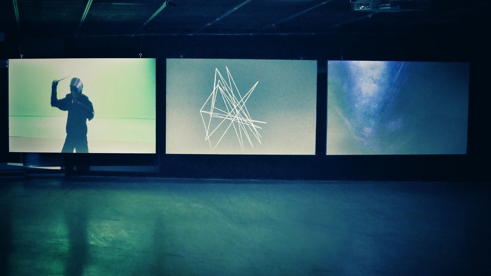 Na zdjęciu ukazane jest pomieszczenie z trzema fotografiami. Na zdjęciu po lewej stronie przedstawiono mężczyznę, który w  prawej ręce trzyma przedmiot, przypominający batutę lub pędzel. Mężczyzna ubrany jest na czarno. Druga fotografia przedstawia obraz z żółtymi liniami na niebieskim tle. Kolejne zdjęcie przedstawia planszę pokrytą odcieniami koloru niebieskiego.  