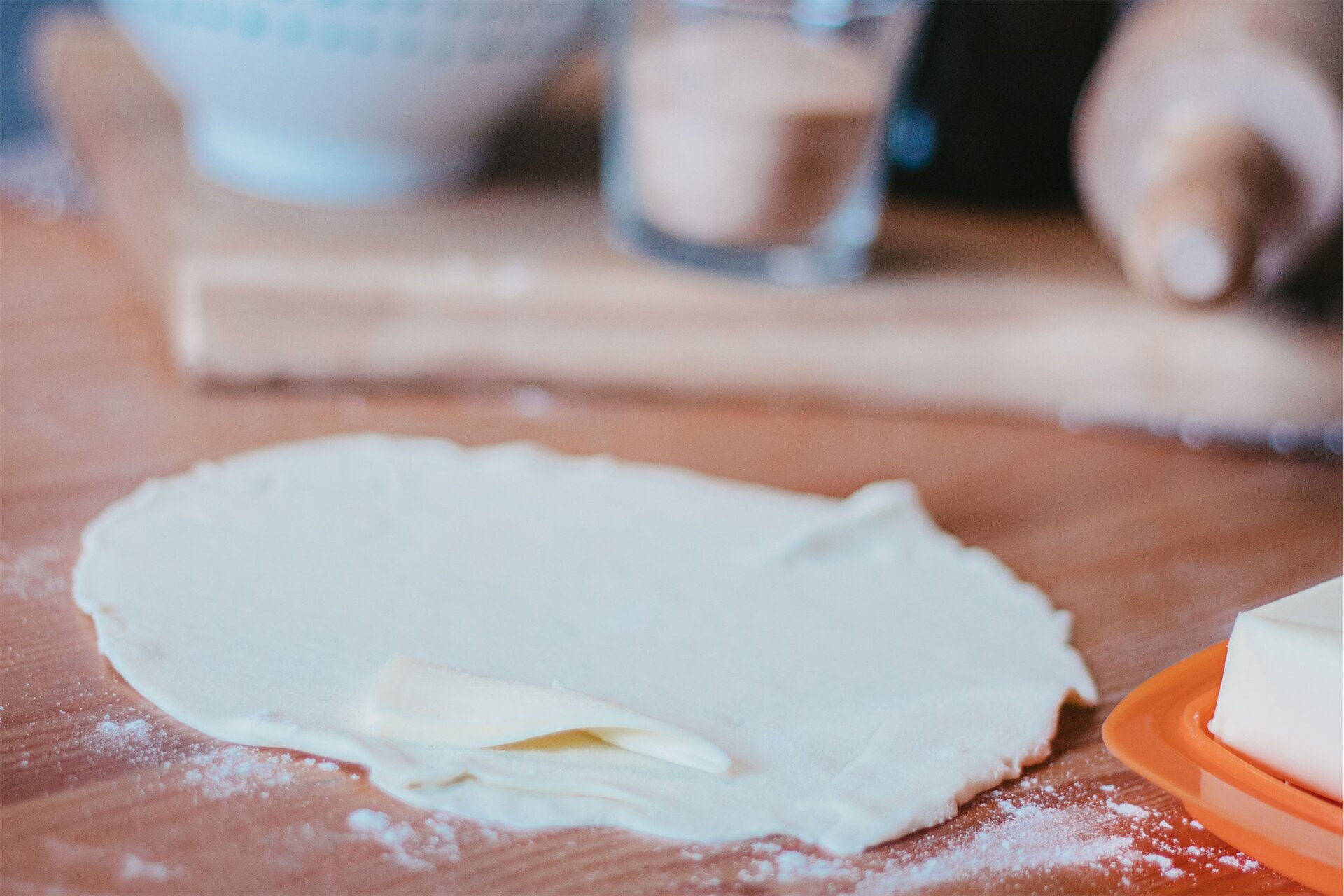 Zdjęcie przedstawia kontynuację kroku drugiego formowania serduszek. Na pierwszym planie widoczny jest cienki owalny płat ciasta oprószony mąką. Na cieście w dolnej części zdjęcia znajduje się kawałek masła w kolorze jasnożółtym. Dookoła płatu ciasta rozypana jest białą mąka. W prawym dolnym rogu grafiki dostrzec można pomarańczową prostokątna maselnicę w środku której znajduje się jasnożółta kostka masła. Na drugim planie widoczny jest niewyraźny zarys drewnianej deski na której znajduje się biała miseczka, szklanka wypełniona cukrem cynamonowym oraz drewniany wałek.