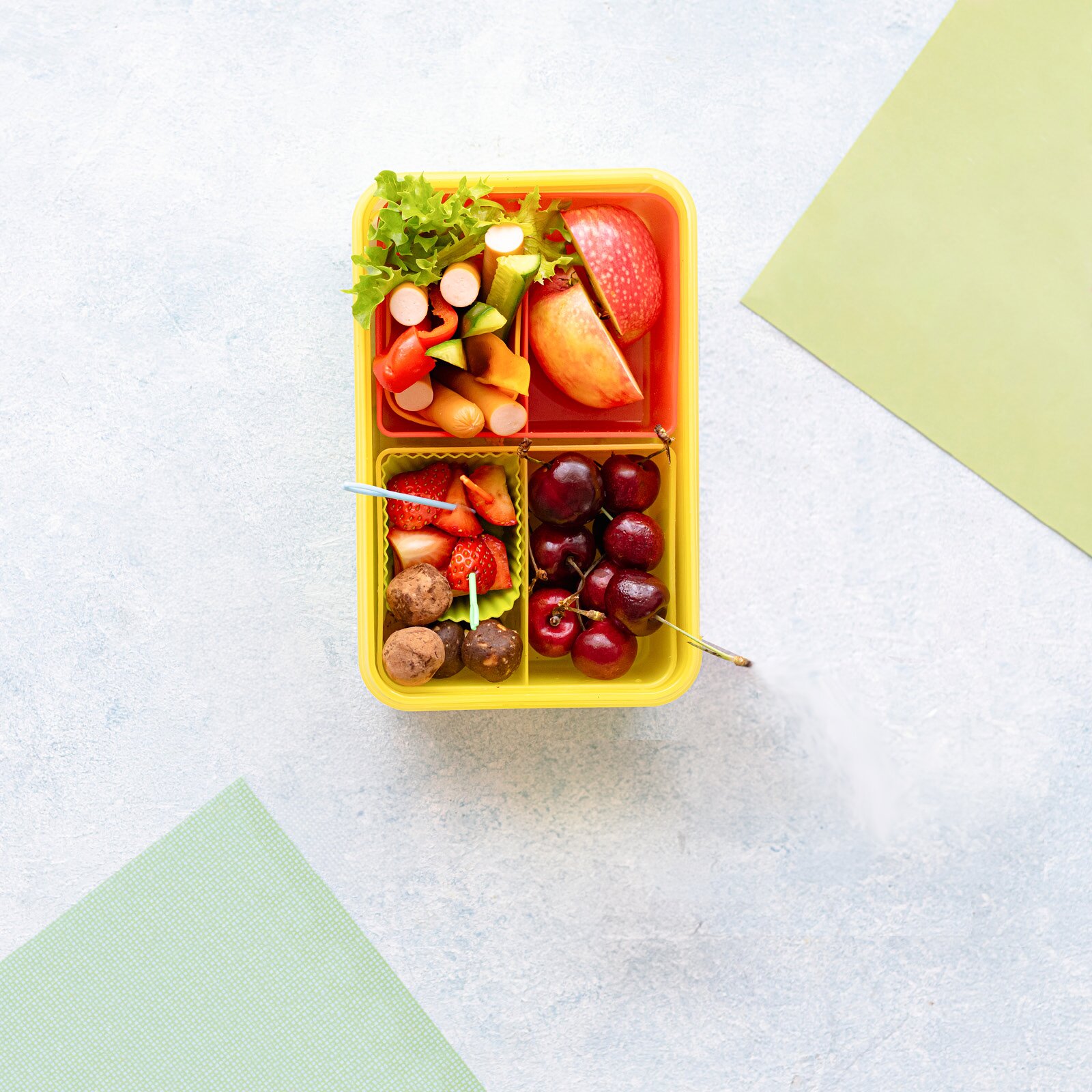 Ilustracja przedstawia pudełko śniadaniowe z czterema przegródkami. W pierwszej przegródce znajduje się rabarbar. W drugiej przegródce znajduje się jabłko przykrojone na pół. W trzeciej przegródce znajdują się truskawki i czekoladki. W czwartej przegródce znajdują się wiśnie.