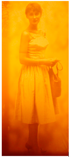 Zdjęcie przedstawia stojącą młodą kobietę. Ubrana jest w jasną sukienkę, trzyma w rękach torebkę, ma założone obcasy. Ma krótkie włosy i pociągłą twarz.