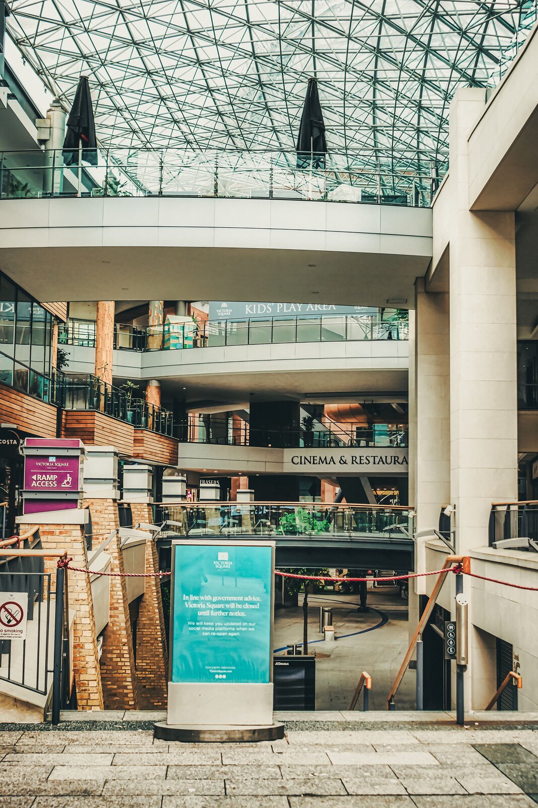 Zdjęcie przedstawia puste wnętrze centrum handlowego z przeszklonym dachem. Widoczne są cztery kondygnacje. Przy schodach prowadzących z najniższej kondygnacji do góry widoczna jest czerwona lina zagradzająca przejście oraz niebieski panel informacyjny z napisem w języku angielskim: VICTORIA SQUARE. In line with government advice, Victoria Square will be closed until further notice. We will keep you updated on our social media platforms, when we can re-open again. W tle widoczne są przeszklone lokale, nad którymi widoczne są napisy w języku angielskim: CINEMA &amp; RESTAURANT, KID’S PLAY AREA.