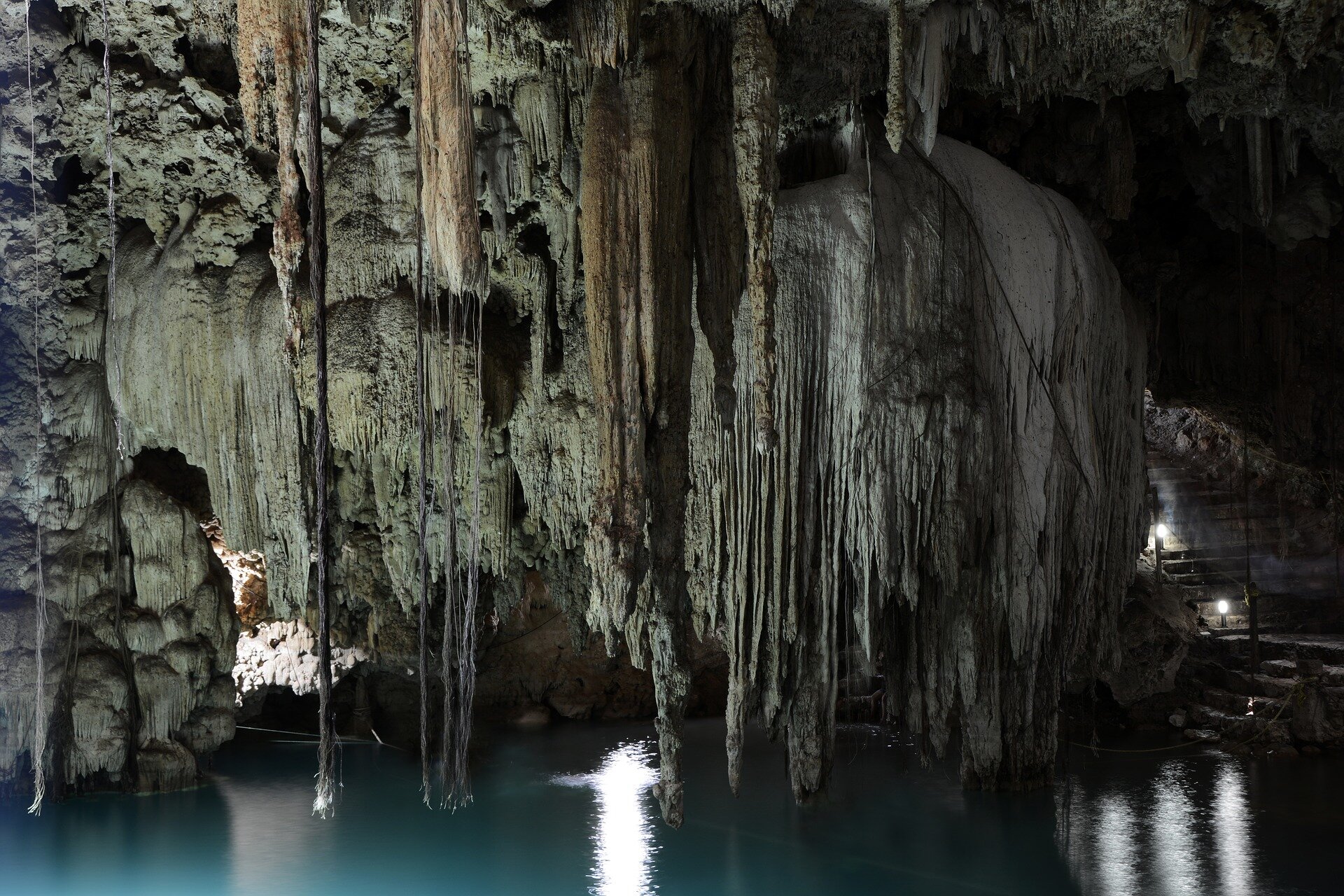 Zdjęcie przedstawiające stalaktyty które zwisają z sufitu jaskini. Stalaktyty wiszą nad wodą, a ich kształt przypomina cienkie rurki. Niektóre są połączone ze sobą i tworzą większe formy - stożki, przypominające sople lodu.