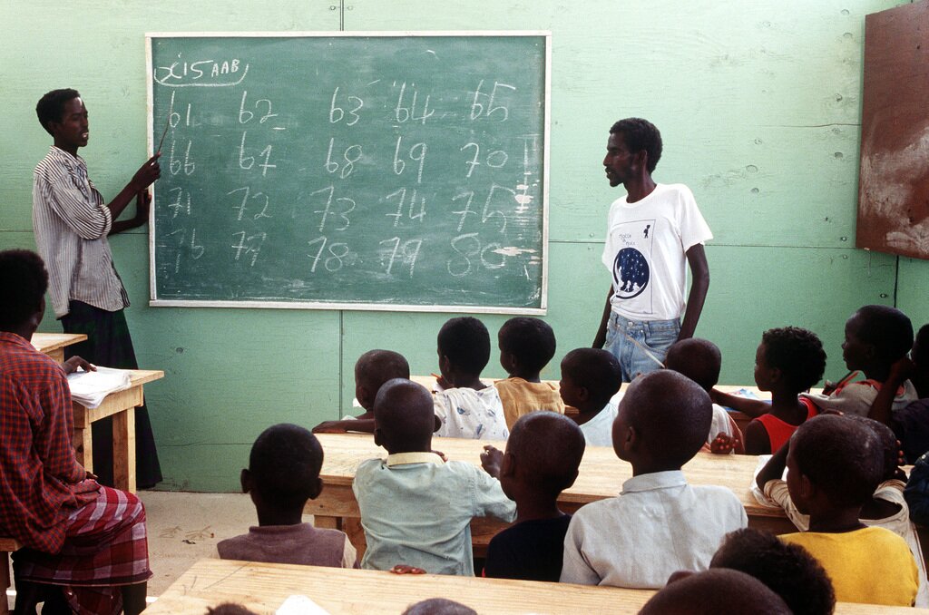 Zdjęcie przedstawia zajęcia w szkole w Somalii. Z prawej strony siedzą w ławkach uczniowie. Jeden z nich stoi przy tablicy. Z lewej strony stoi nauczyciel pokazujący ręką na tablicę, na której znajdują się wypisane liczby.