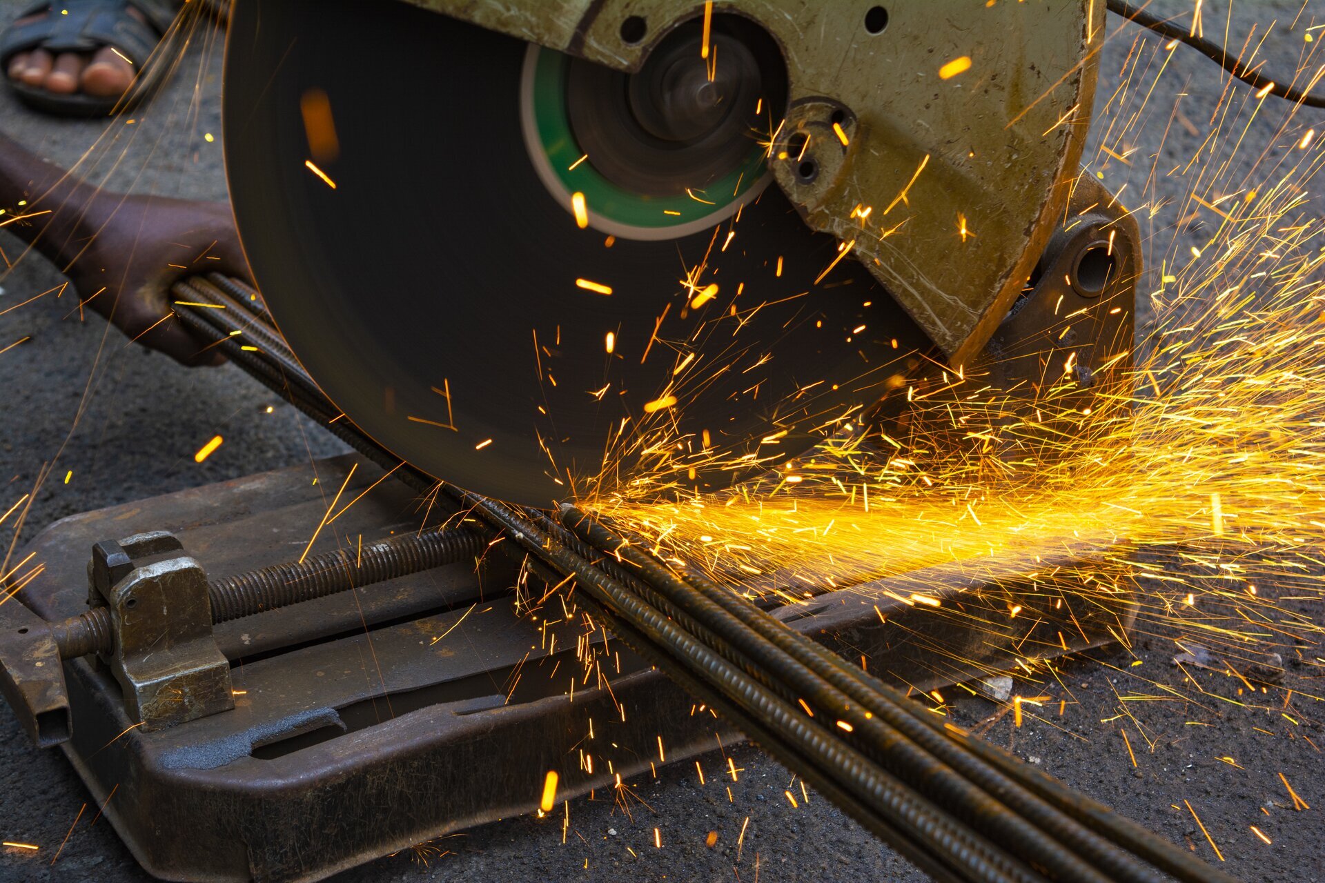 Zdjęcie przedstawia szlifierkę kątową o dużej średnicy ostrza, którą postać niewidoczna na zdjęciu przecina stalowy pręt.