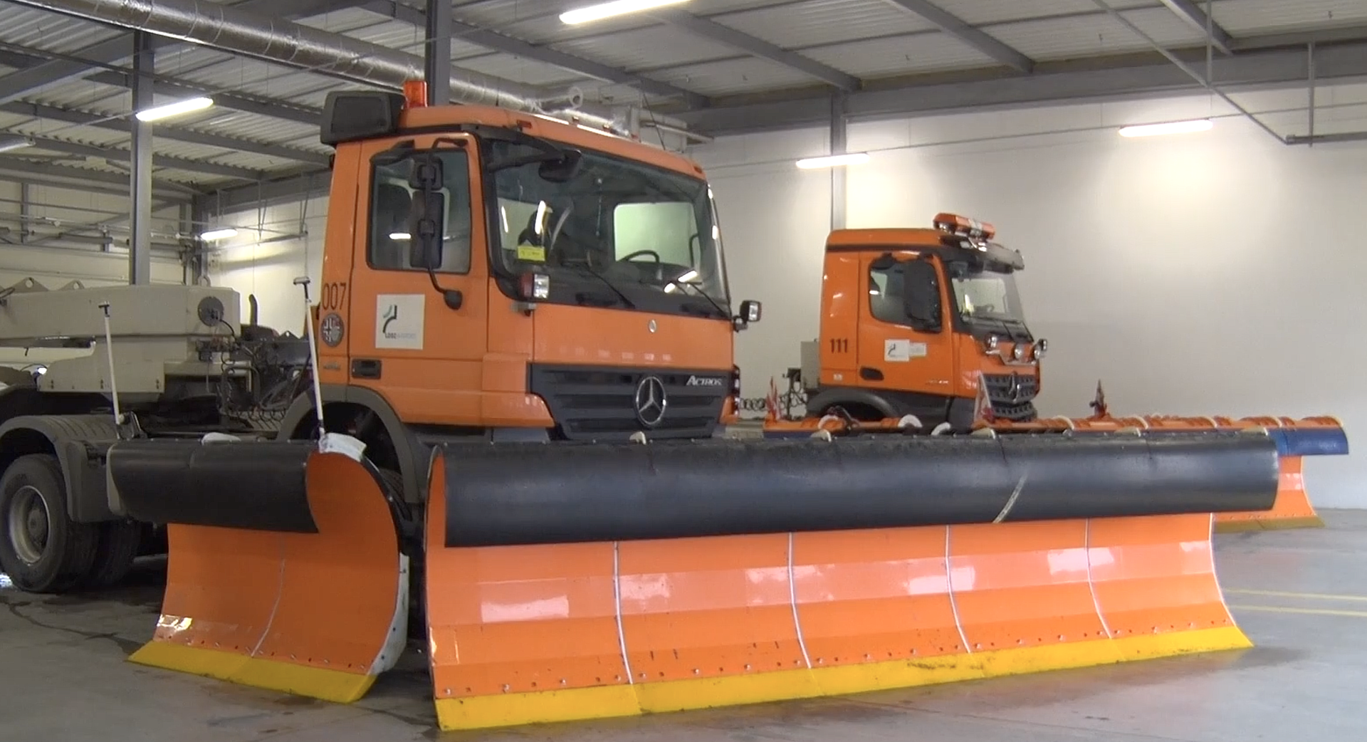 Zdjęcie przedstawia pług odśnieżający płytę lotniskową. Jest to duży ciężki pojazd, który z przodu ma zamontowaną odpowiednio wygiętą metalową część. Służy ona do zgarniania śniegu z powierzchni płyty. Jest szeroka i niska i w przybliżeniu ma prostokątny kształt.