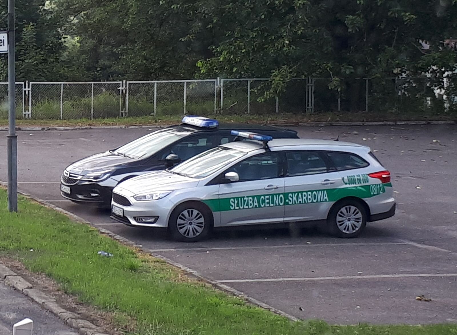 Na zdjęciu pojazd Służby Celno‑Skarbowej — srebrne combi. Na bocznych drzwiach zielony pas z napisem "Służba Celno‑Skarbowa". Na końcu zielonego paska, znajduje się numer ewidencyjny samochodu, w tym przypadku to 08/12. Nad zielonym paskiem, na wysokości bagażnika znajduje się zielony numer alarmowy 0 800 060 000.  Na dachu znajdują się  niebieskie sygnalizatory świetlne. Obok samochodu służby skarbowej stoi drugi, prawie cały przysłonięty, ciemnogranatowy samochód combi. Samochody stoją na parkingu.