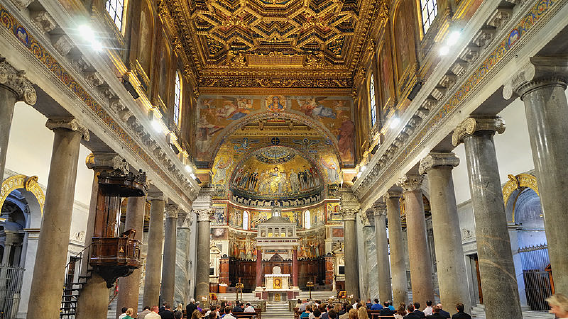 Zdjęcie przedstawia wnętrze bazyliki Najświętszej Maryi Panny na Zatybrzu w Rzymie. Na zdjęciu widoczna jest główna nawa bazyliki i prezbiterium. Widoczne są freski i zdobienia na suficie. W ławkach siedzą ludzie.