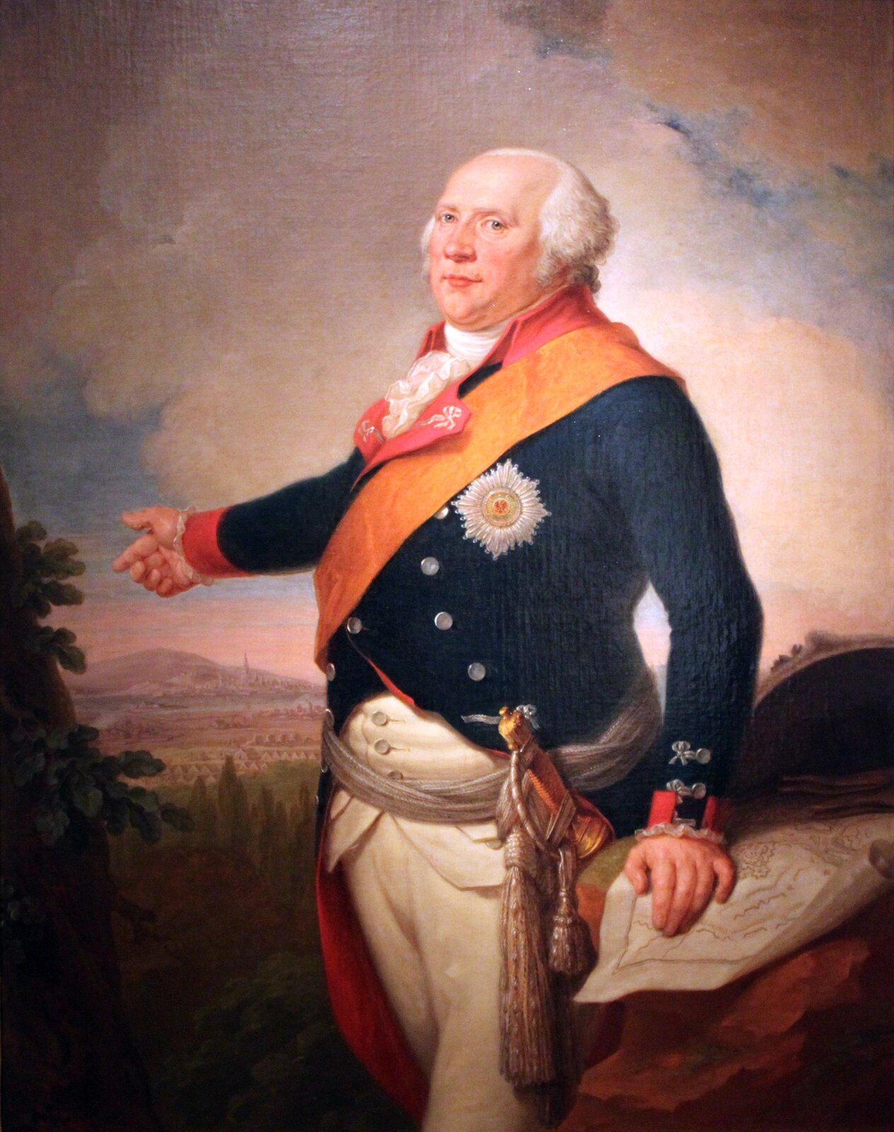 Obraz przedstawia starszego mężczyznę z dużymi zakolami. Ubrany jest w białą koszulę, niebiesko‑czerwoną marynarkę oraz białe spodnie. Przez ramię przewieszoną ma pomarańczową szarfę. W biodrach przewiązany ma szary sznur, do którego przymocowana została szabla. Prawą dłonią wskazuje polanę, za którą widoczne jest miasto i góra, a lewą dłoń opiera o mebel.