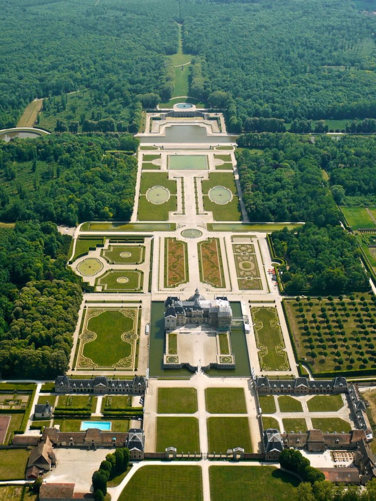 Zdjęcie przedstawia Kompleks pałacowo‑ogrodowy Vaux‑le‑Vicomte, centralna cześć ogrodu, autorstwa Alain Rouleu. Fotografia zrobiona jest z perspektywy lotu ptaka. Na środku znajduje się pałac. Wokół głównego budynku znajdują się liczne ogrody w kształcie kwadratów, okręgów i prostokątów. W prawym i lewym dolnym rogu znajdują się budynki, należące do kompleksu pałacowego. Pomiędzy nimi znajduje się ozdobna brama wejściowa. Cały kompleks otoczony jest lasami. 