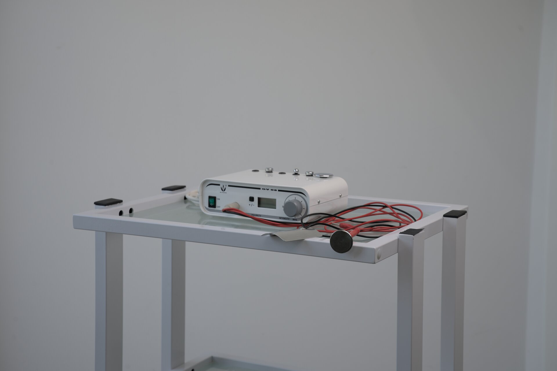 Zdjęcie przedstawia aparaturę do galwanizacji, która stoi na stoliku. Aparatura składa się z urządzenia, które na obudowie posiada niewielki wyświetlacz, przyciski oraz pokrętło. Do urządzenia za pomocą czerwonego i czarnego przewodu podłączone są dwie elektrody. Jedna z nich jest płaska a druga okrągła.