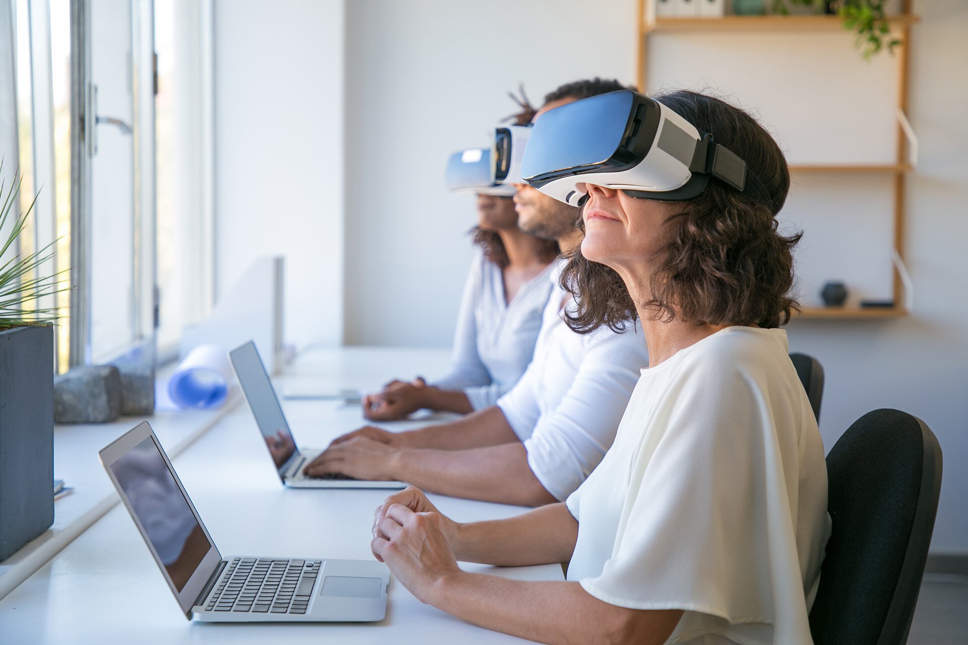 Zdjęcie przedstawia trzy osoby: kobietę, mężczyznę i kobietę, którzy siedzą obok siebie przy długim biurku. Na blacie przed nimi stoją otwarte laptopy. Każda z osób ma na głowie założone gogle do rozszerzonej rzeczywistości. Gogle mają grubość około 10 cm, a utrzymuje je opaska nasunięta na tył głowy. 