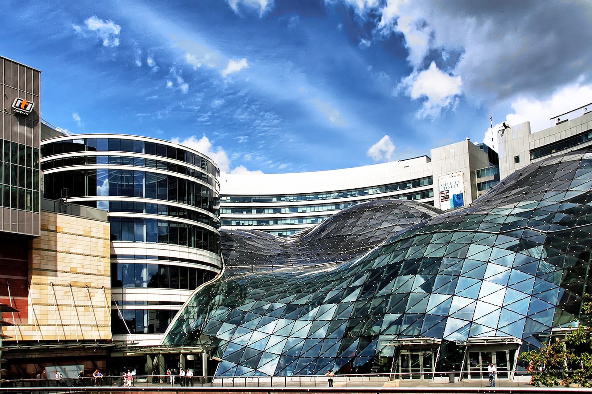 Ilustracja o kształcie poziomego prostokąta przedstawia Złote tarasy. zaprojektowane przez Jerde Partnershipa. Fotografia ukazuje widok od strony placu na wejście frontalne do obiektu wraz z otoczeniem architektonicznym. Na uwagę zasługuje widoczne szklany dach, którego organiczny, falisty kształt został zaprojektowany tak, aby przypominać starożytne zadaszenia koronujące parki warszawskie. Pofalowana forma dachu tworzy wciągające i wysoce zindywidualizowane przestrzenie poruszające się po tarasach. Po lewej stronie widoczne są inne obiekty handlowo-przemysłowe. 