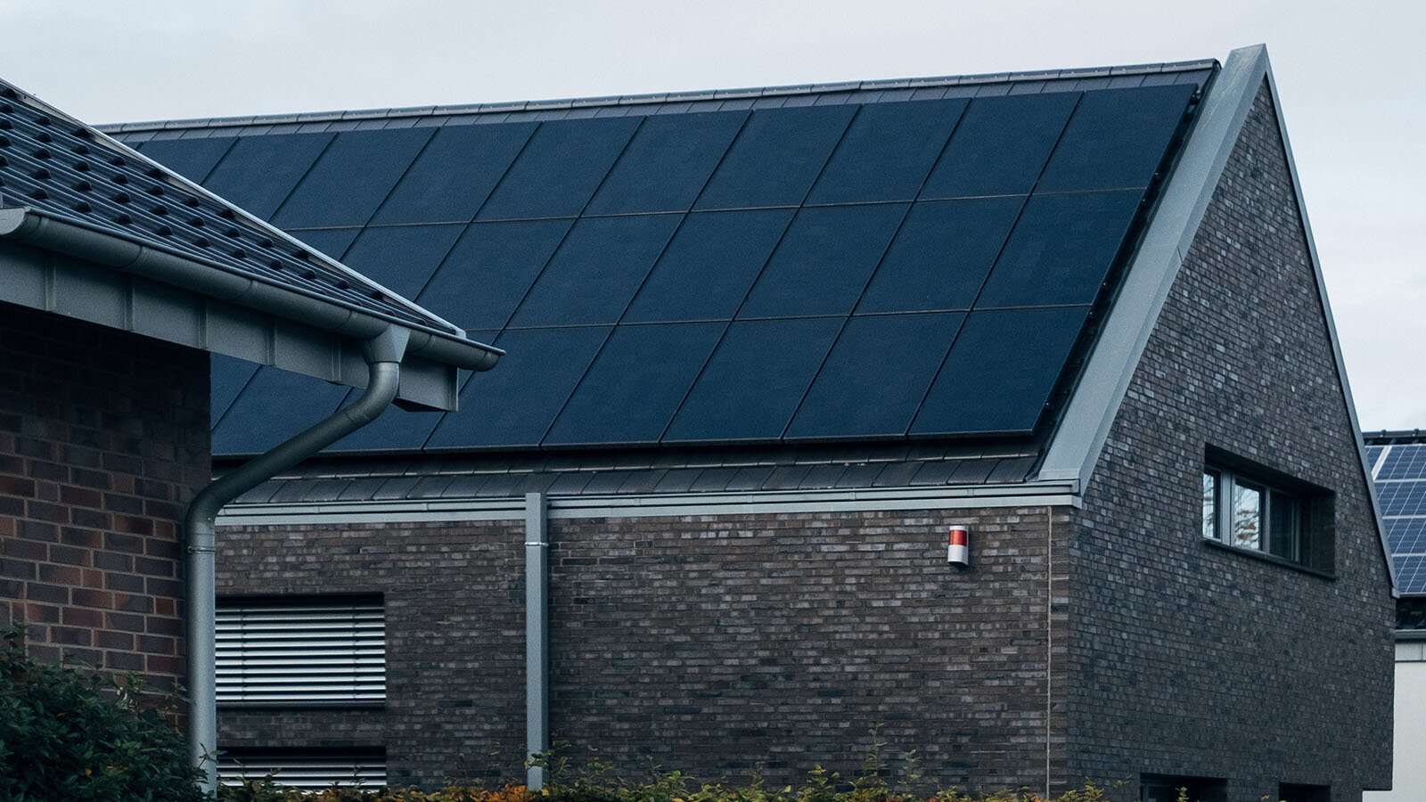 Zdjęcie przedstawia dom, którego dach pokrywają panele słoneczne. Elewacja domu wykonana jest z ciemnej płytki klinkierowej. 