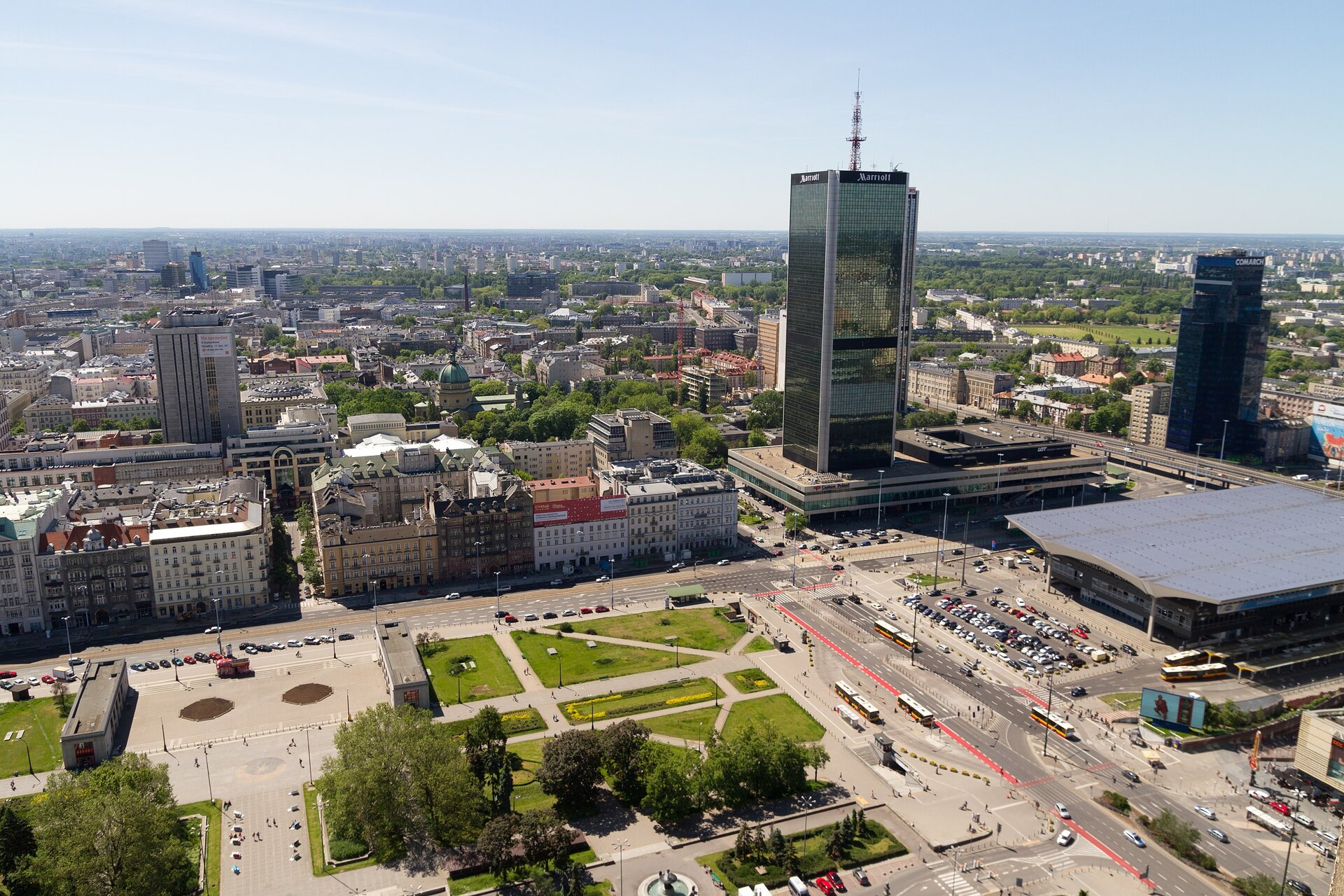 Zdjęcie przedstawia panoramę miasta. Na pierwszym planie znajduje się plac, który częściowo pokryty jest trawą. Po prawej stronie jest się niewysoki, płaski budynek dworca. Obok niego stoi wysoki wieżowiec hotelu. Po lewej stronie znajdują się niewysokie kamienice.