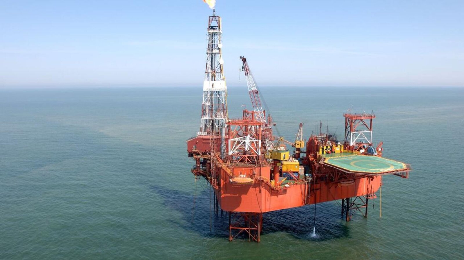 Platforma wiertnicza stojąca na otwartym morzu. Ma czerwony kolor, konstrukcja wznosi się powyżej linii wody na metalowych filarach. Z lewej strony wieża z dużym płomieniem na końcu, z prawej lądowisko dla helikoptera.