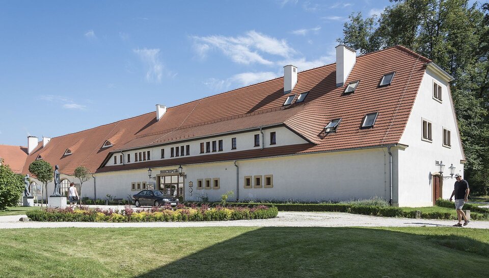 Na zdjęciu znajduje się duży podłużny budynek o stromym czerwonym dachu i białych ścianach. Posiada małe okna i kilka kominów. Przed wejściem na dróżce stoi współczesny samochód. Przed budynkiem znajdują się pomniki i park. Za budynkiem rosną drzewa. 